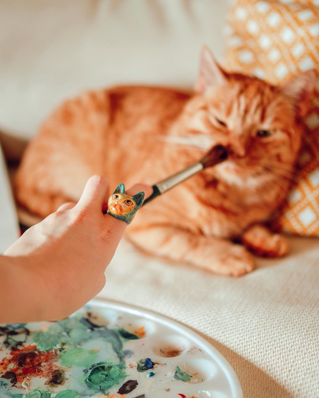 Hand holding a cat-shaped ring near a sleepy orange cat on a couch, with a paint palette in the foreground.