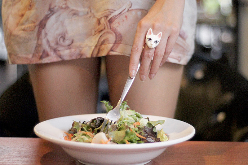 A hand with a cute white cat ring holds a fork over a fresh salad in a bowl.