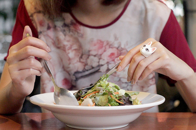 A hand with a cute white cat ring holds a fork and spoon over a fresh salad in a bowl.