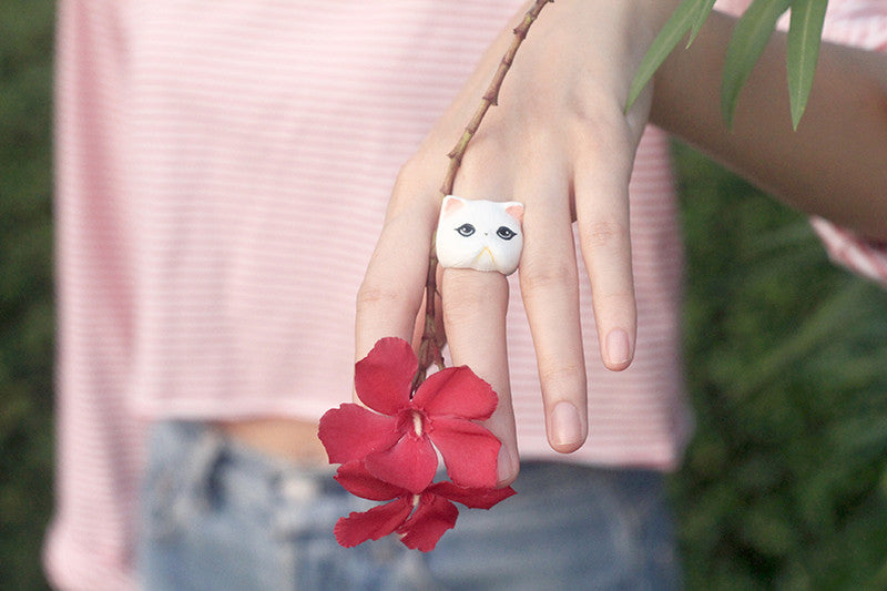 A cute cat face ring worn on a hand next to a red flower.