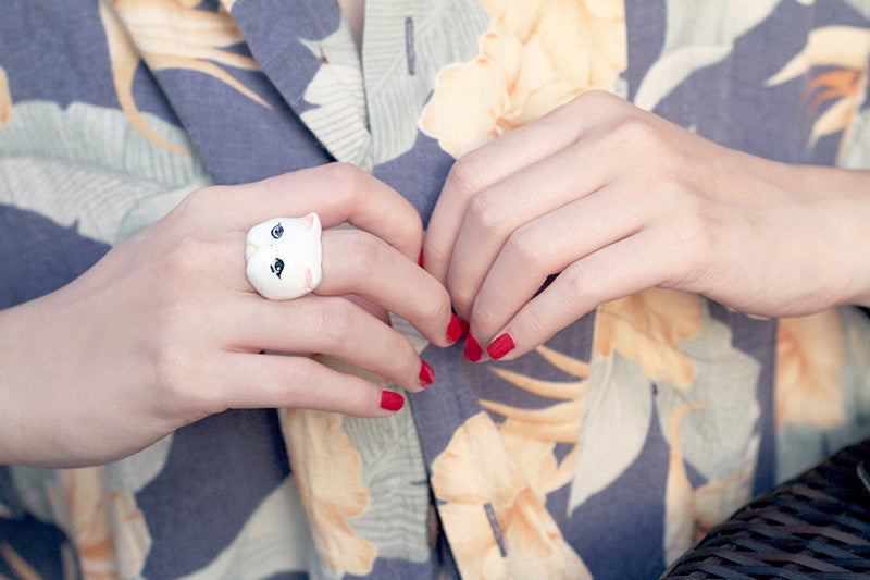 A hand with a white cat ring and red nails, dressed in a floral shirt.