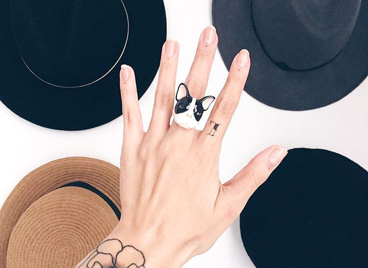 Close-up of a cute French Bulldog ring on a hand, with hats in the background.