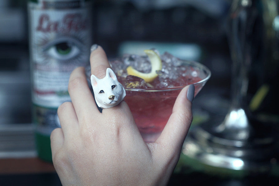 A close-up of a hand adorned with a white Siberian husky ring holding a pink cocktail garnished with lemon.