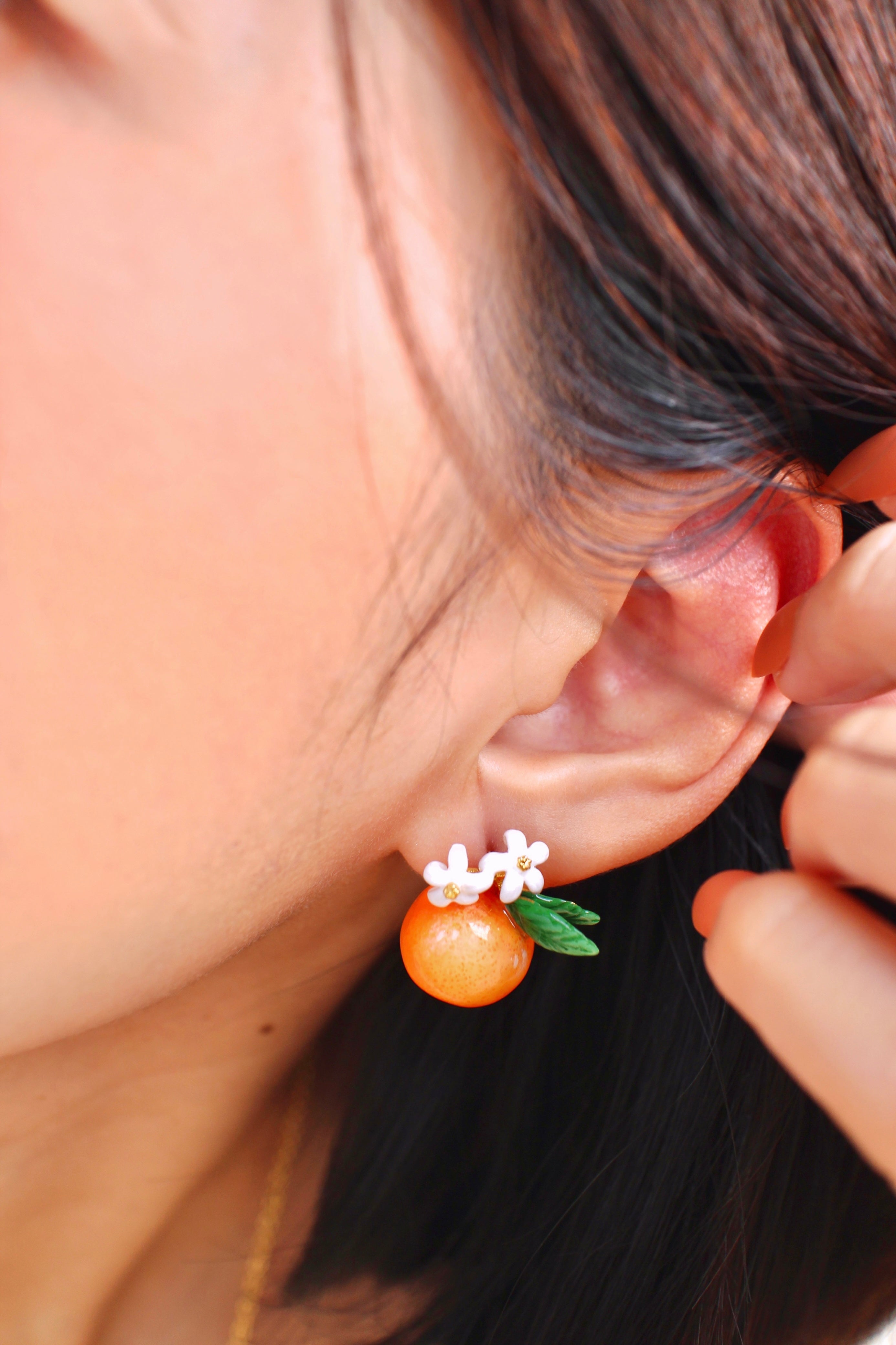 A close-up of an ear wearing charming orange earrings adorned with white flowers and green leaves.