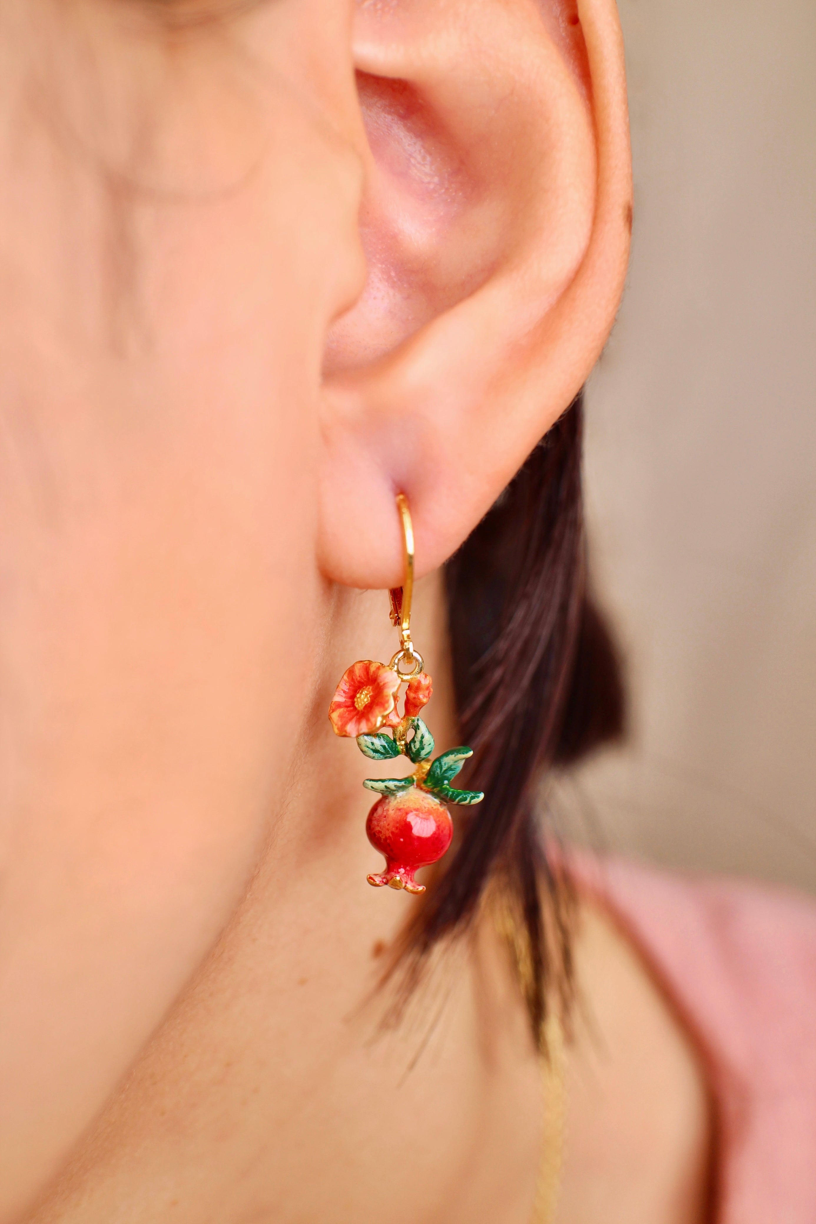 A close-up of an ear wearing charming pomegranate earrings adorned with red flowers and green leaves.