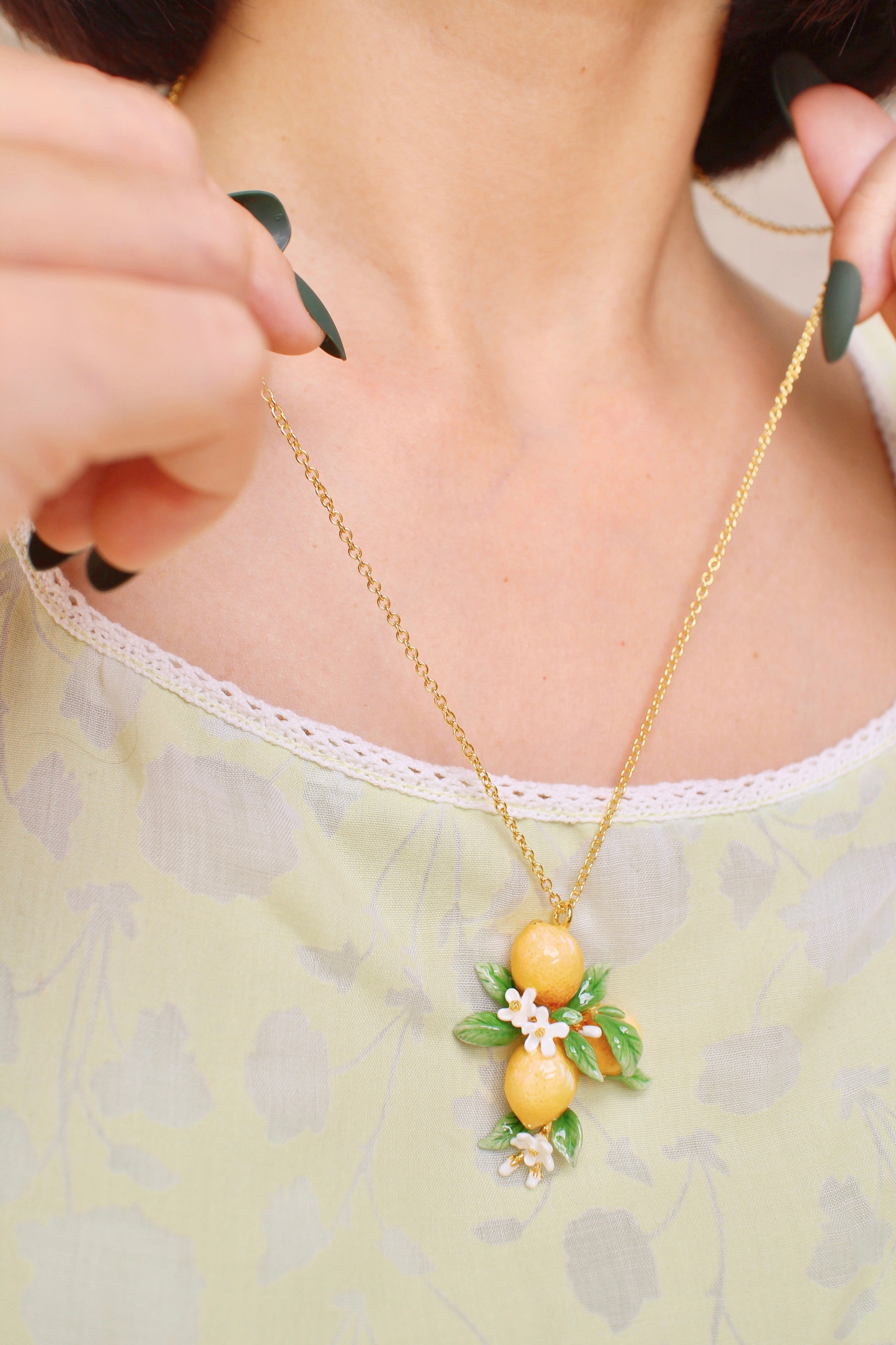 A close-up of a person holding a gold chain necklace with lemon charms and white flower accents, in a summer outfit.