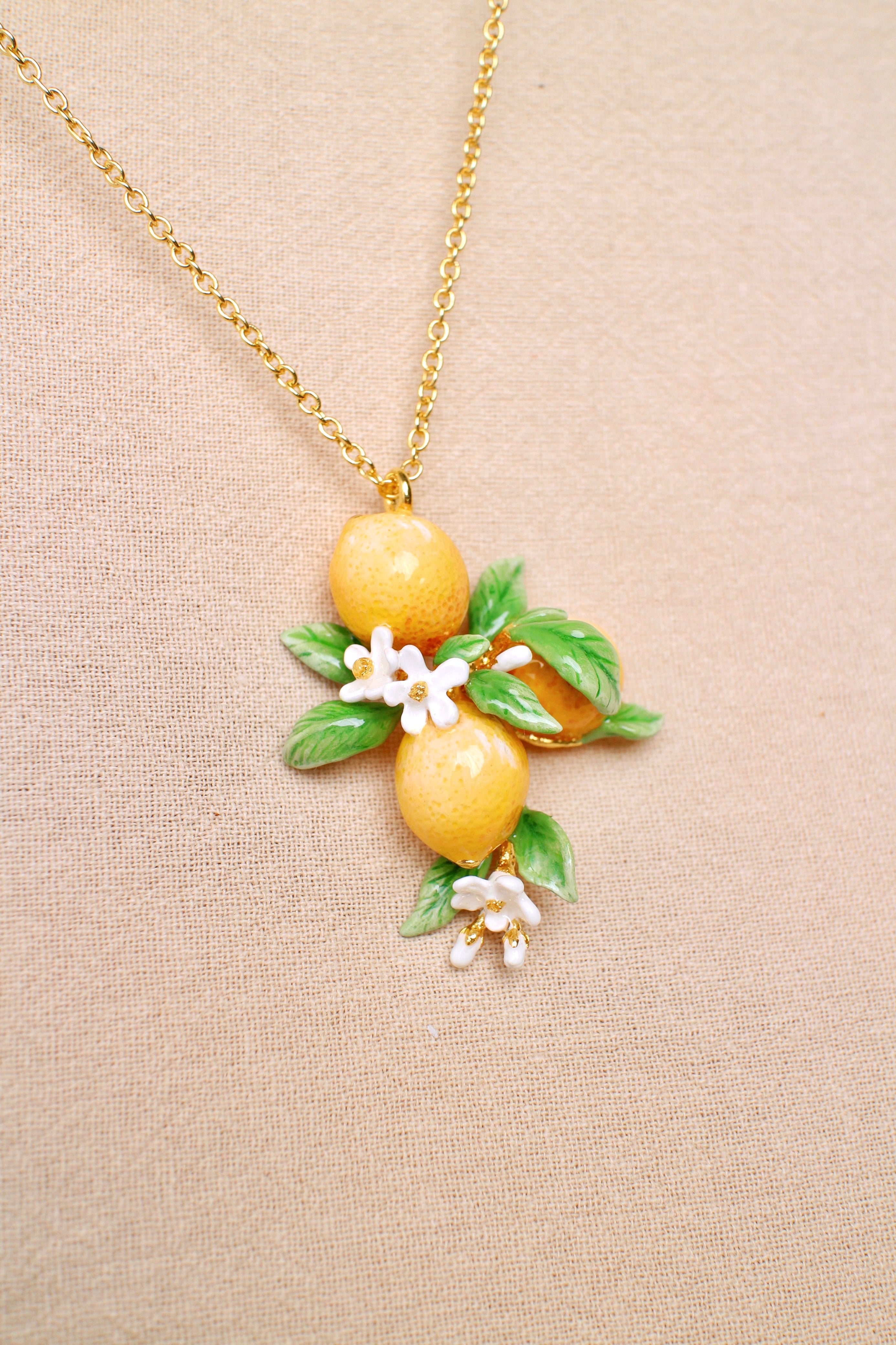 A golden chain necklace featuring three intricately crafted yellow lemons surrounded by green leaves and tiny white flowers.