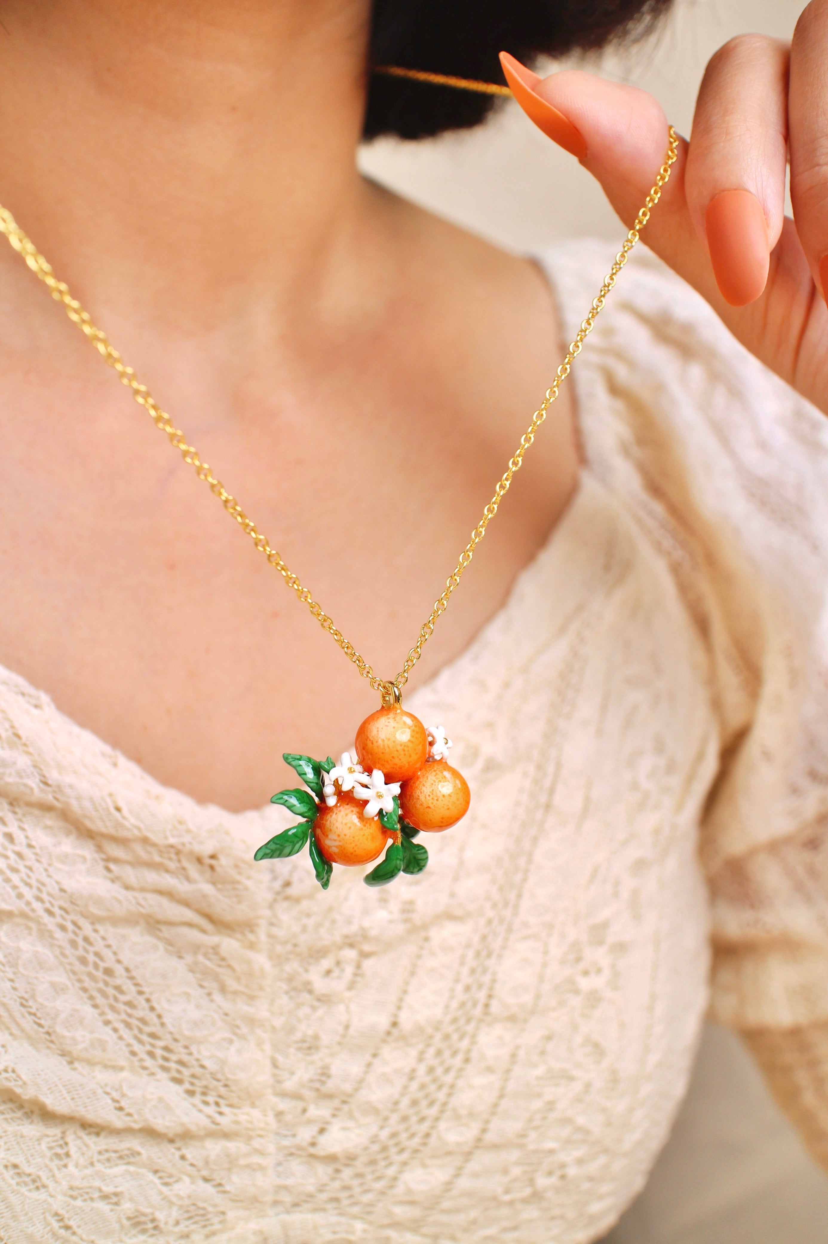 A close-up of a person holding a gold chain necklace with orange charm and white flower accents, in a summer outfit.