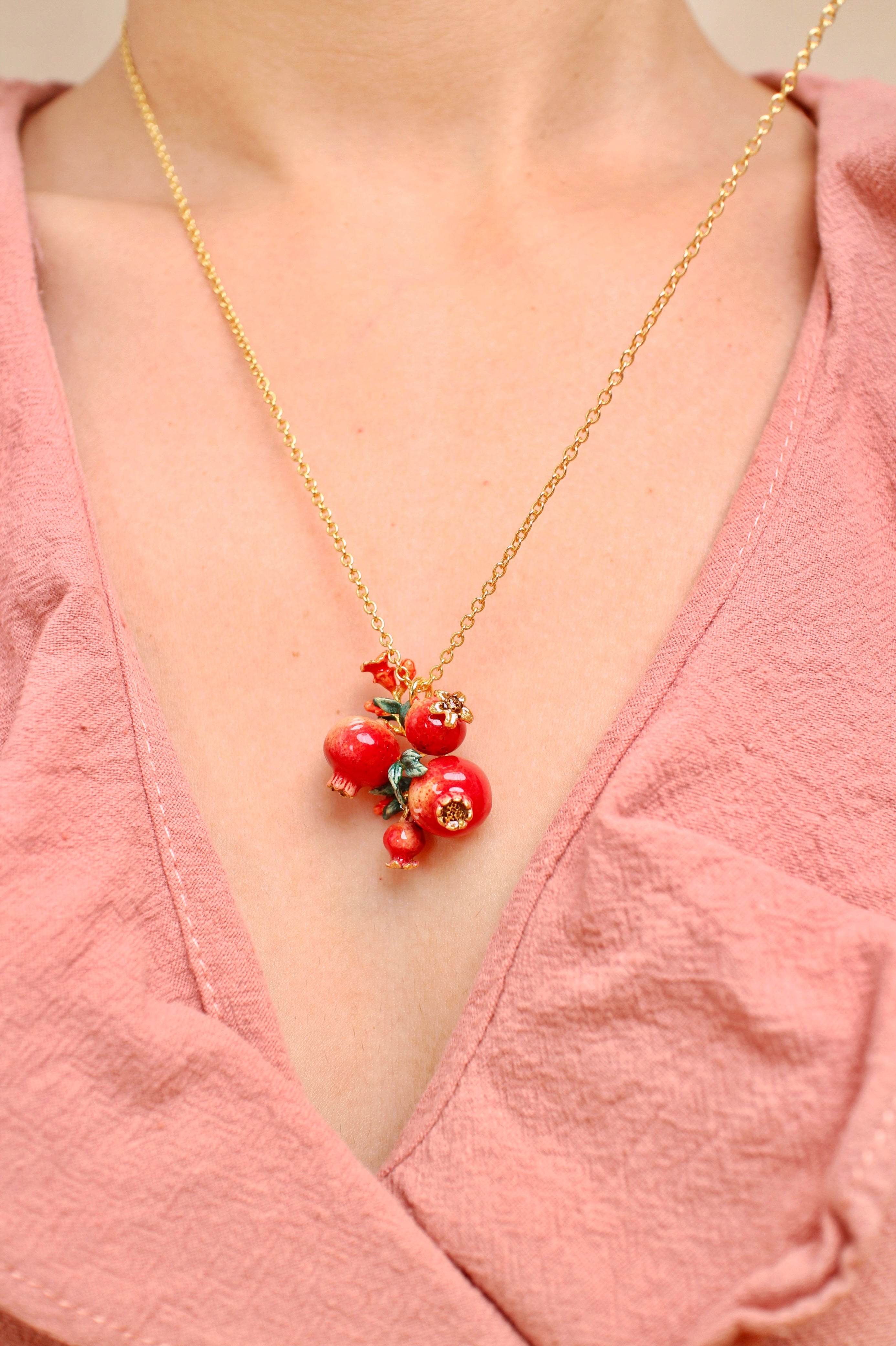 A close-up of a person wearing a gold chain necklace with pomegranate charm and green leaves accents, in a summer outfit.