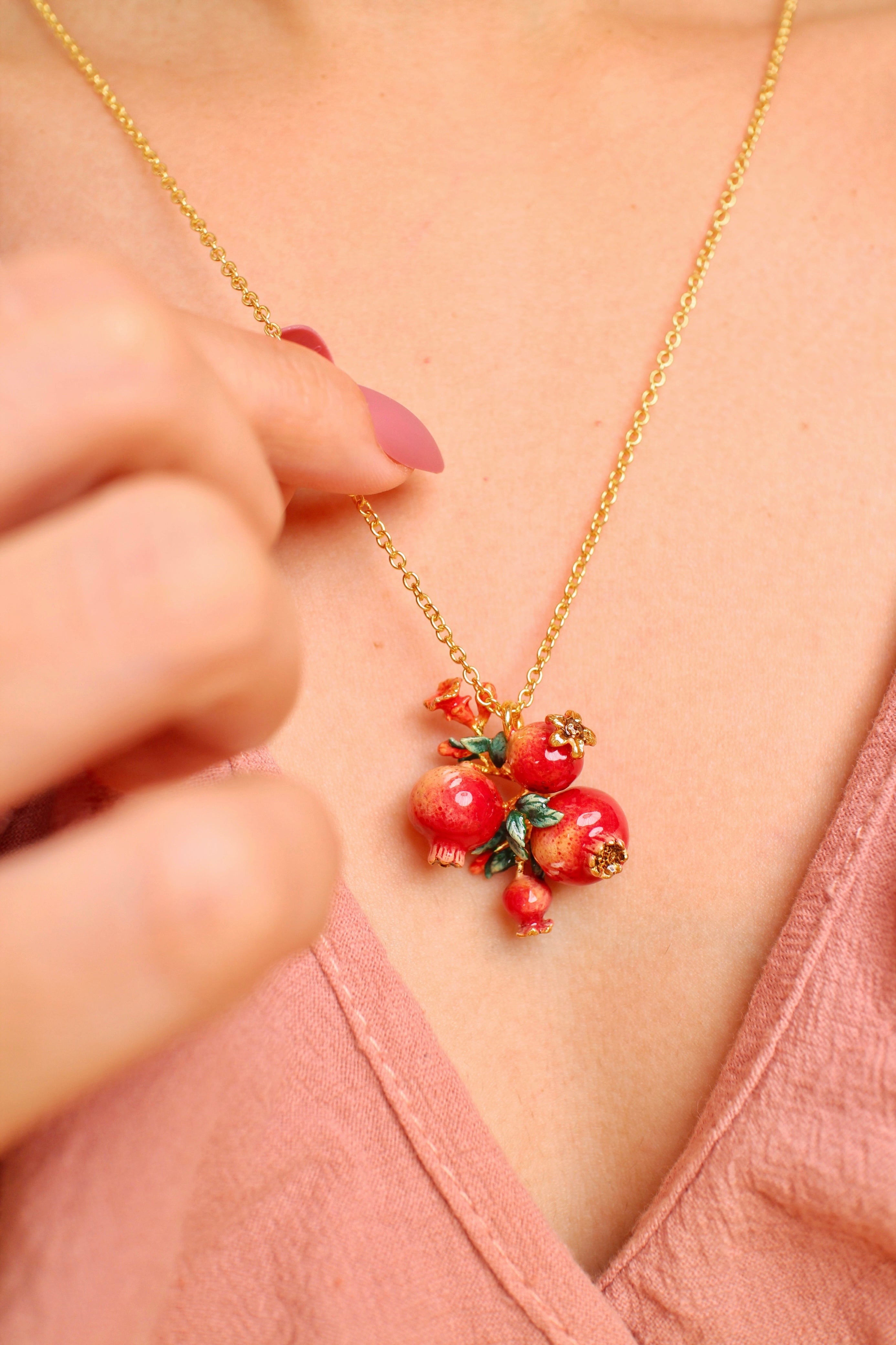 A close-up of a person wearing a gold chain necklace with pomegranate charm and green leaves accents, in a summer outfit.