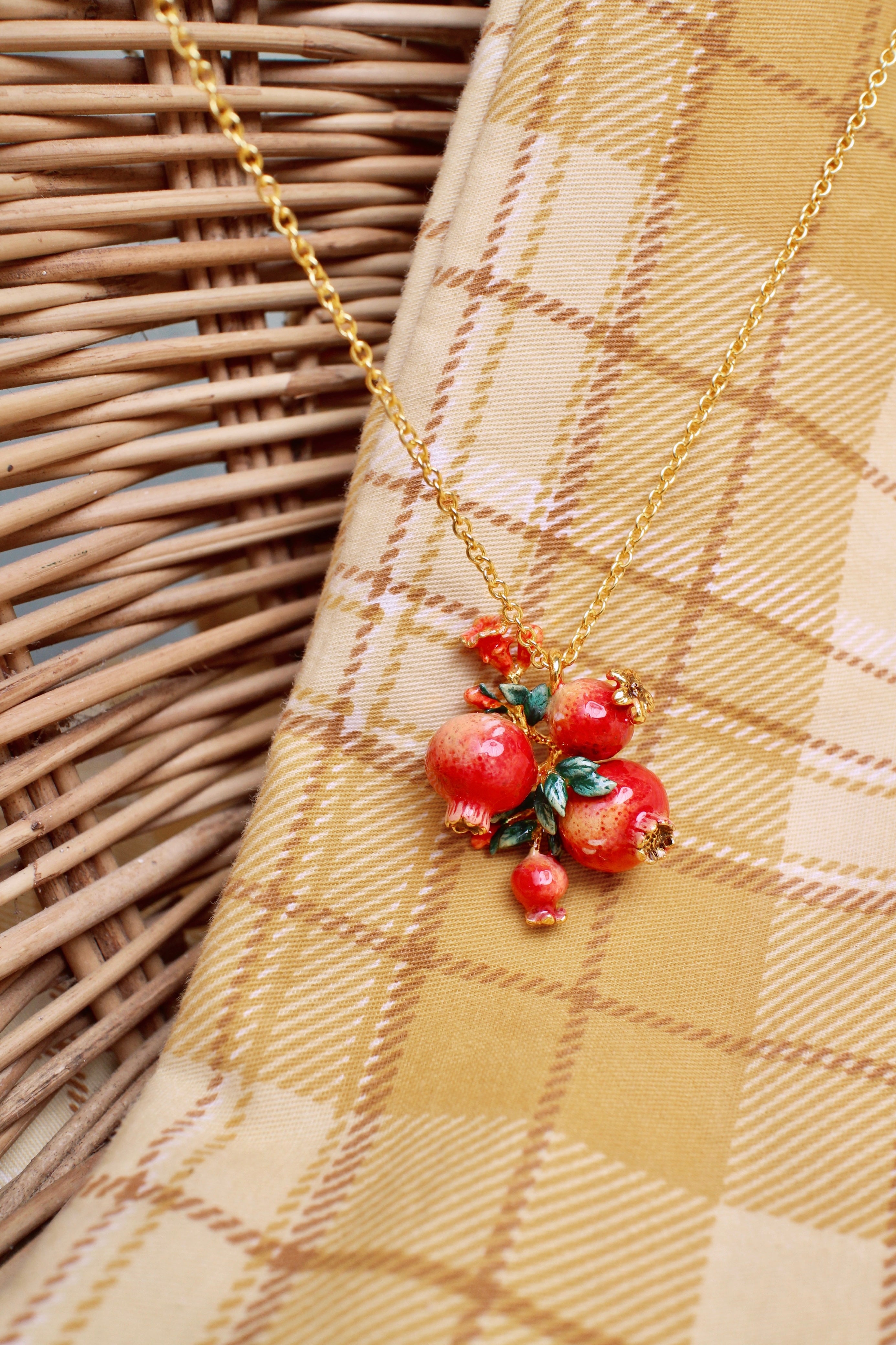 A golden chain necklace featuring three intricately crafted pomegranates surrounded by green leaves and tiny red flowers placed on a fabric and a wicker basket.