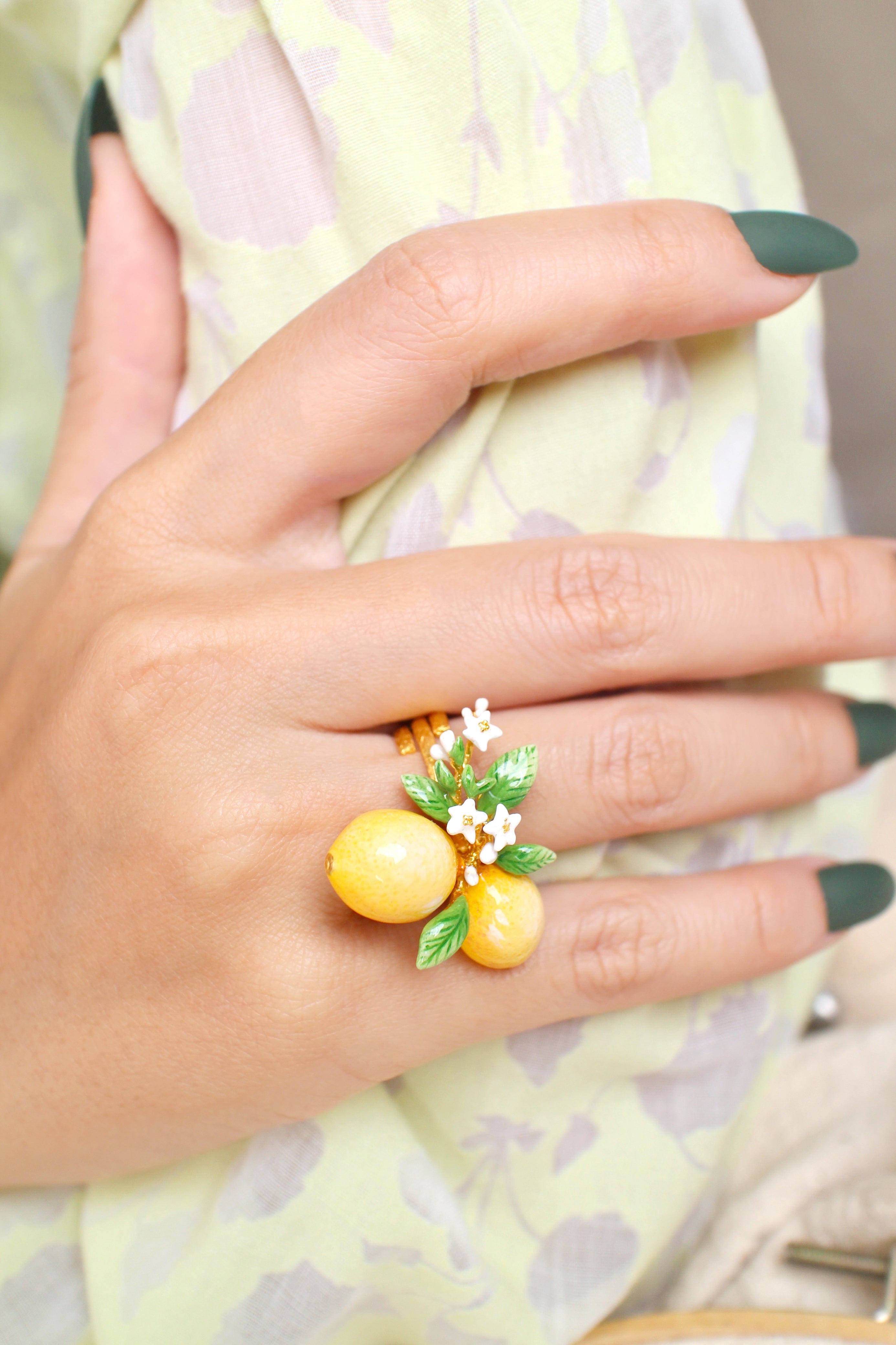 A hand wearing a handmade ring featuring two yellow lemons, green leaves, and small white flowers, in a summer outfit.