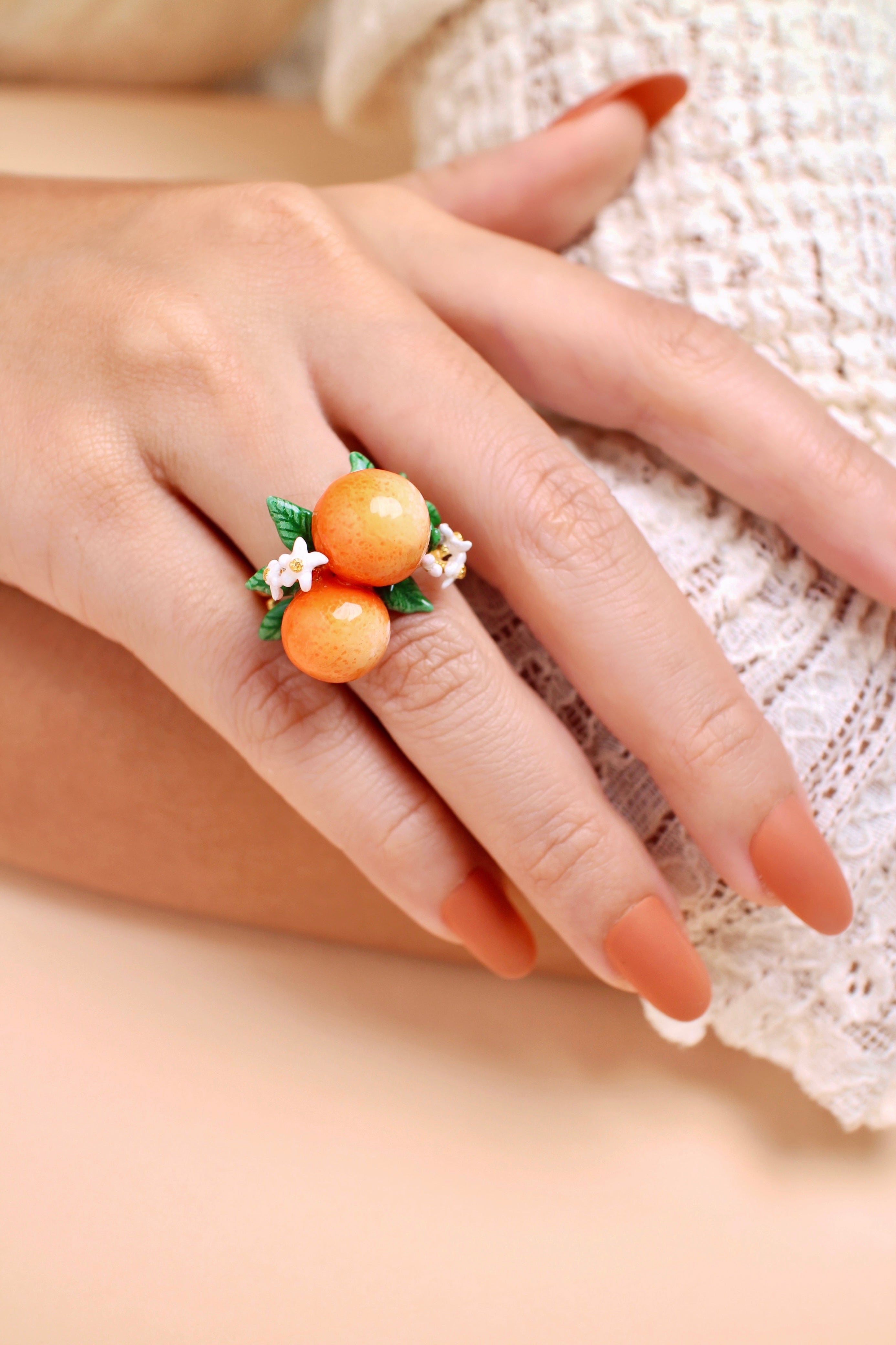 A hand wearing a handmade ring featuring two oranges, green leaves, and small white flowers, in a summer outfit.