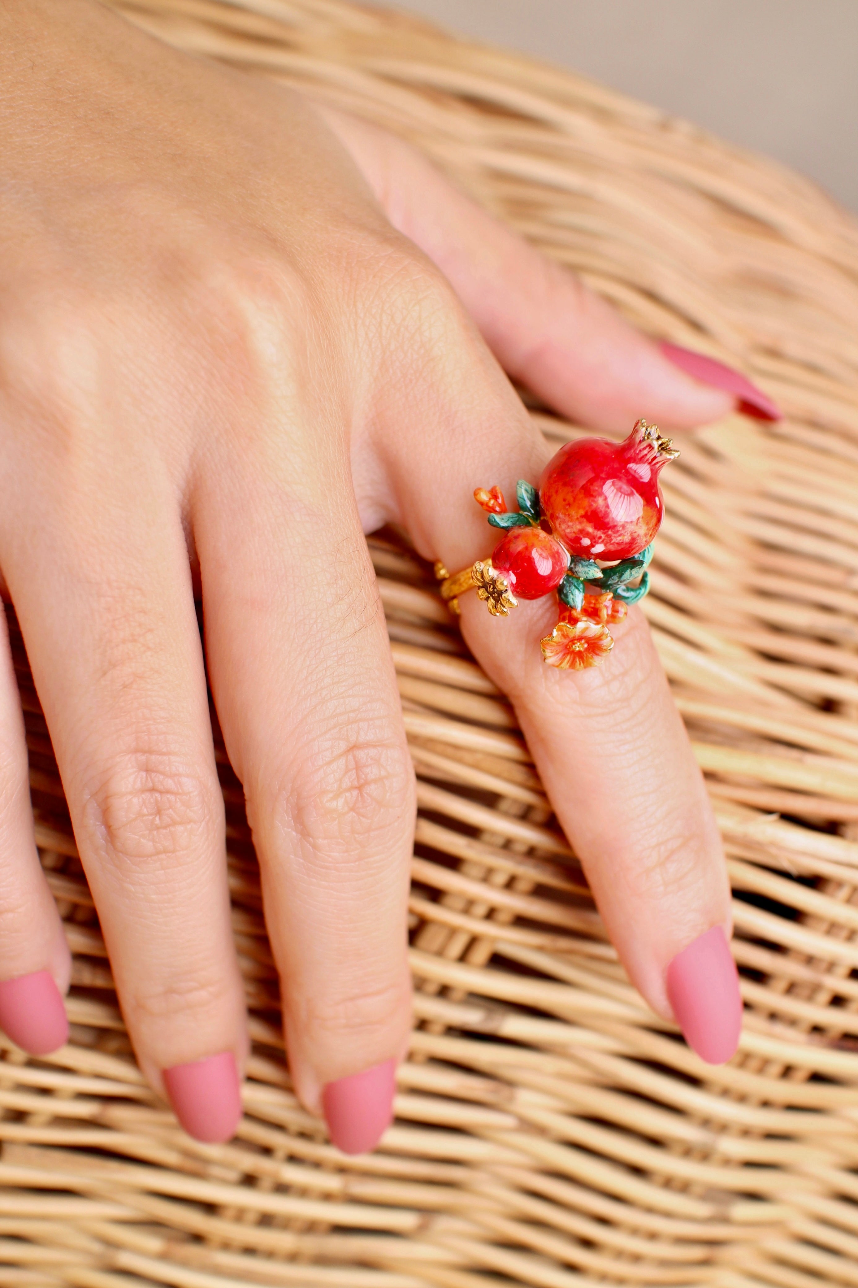 Close-up of a hand wearing a handmade ring, featuring two pomegranates, green leaves, and small red flowers, rests on a wicker basket.