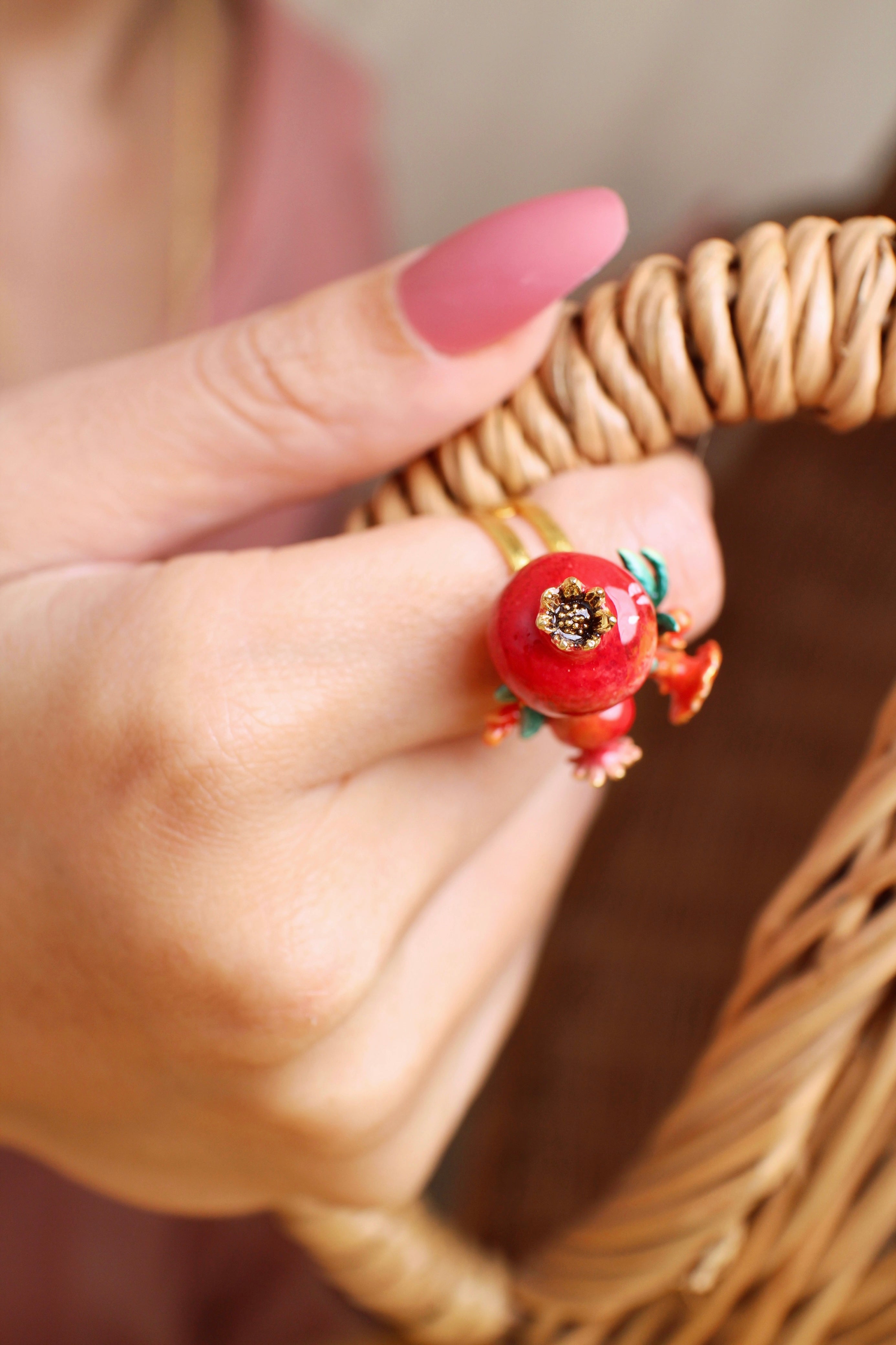 Close-up of a hand wearing a handmade ring, featuring two pomegranates, green leaves, and small red flowers, holding a handle of a wicker basket.