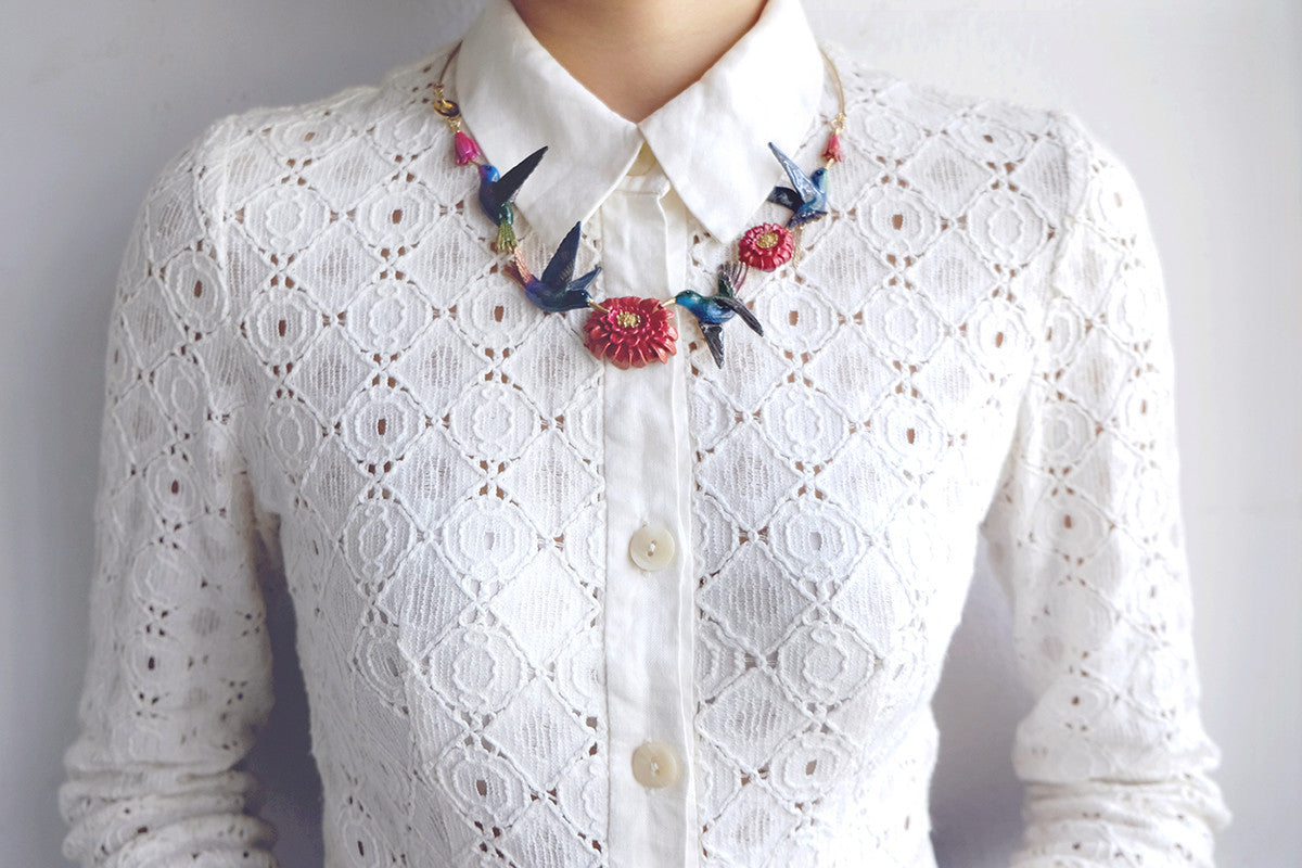 A statement necklace featuring hummingbirds and floral elements, styled with a white lace top.