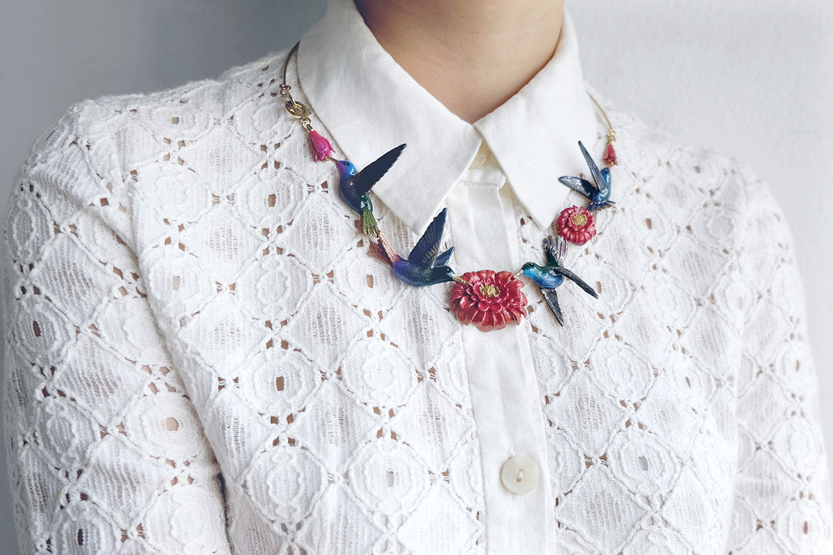 A statement necklace featuring hummingbirds and floral elements, styled with a white lace top.