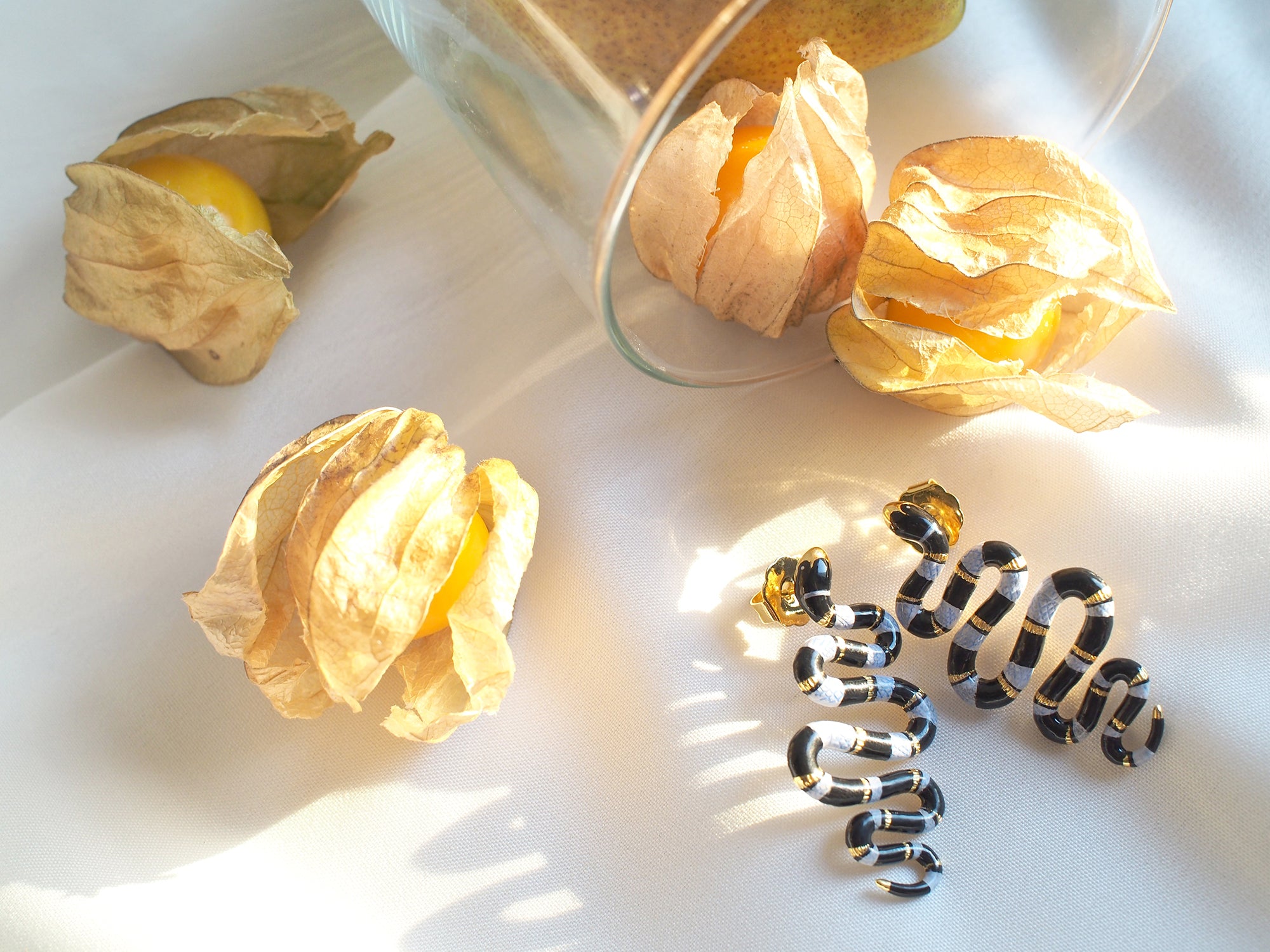 Handmade earrings featuring blue sea snake with a black stripe rest next to gooseberries on a soft pastel background.