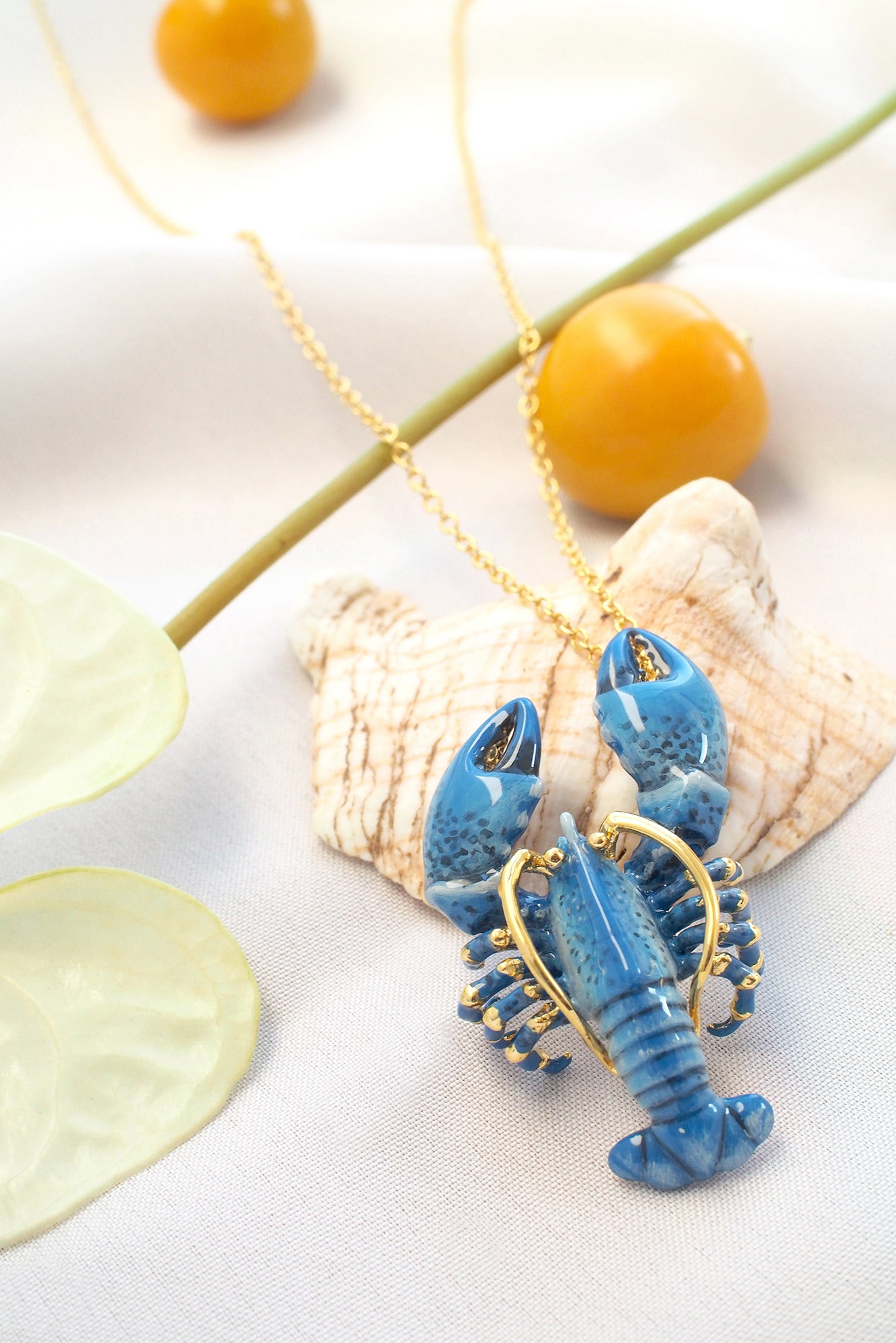 Blue lobster-shaped pendant necklace with gold accents on a delicate chain placed on a seashell and natural element.