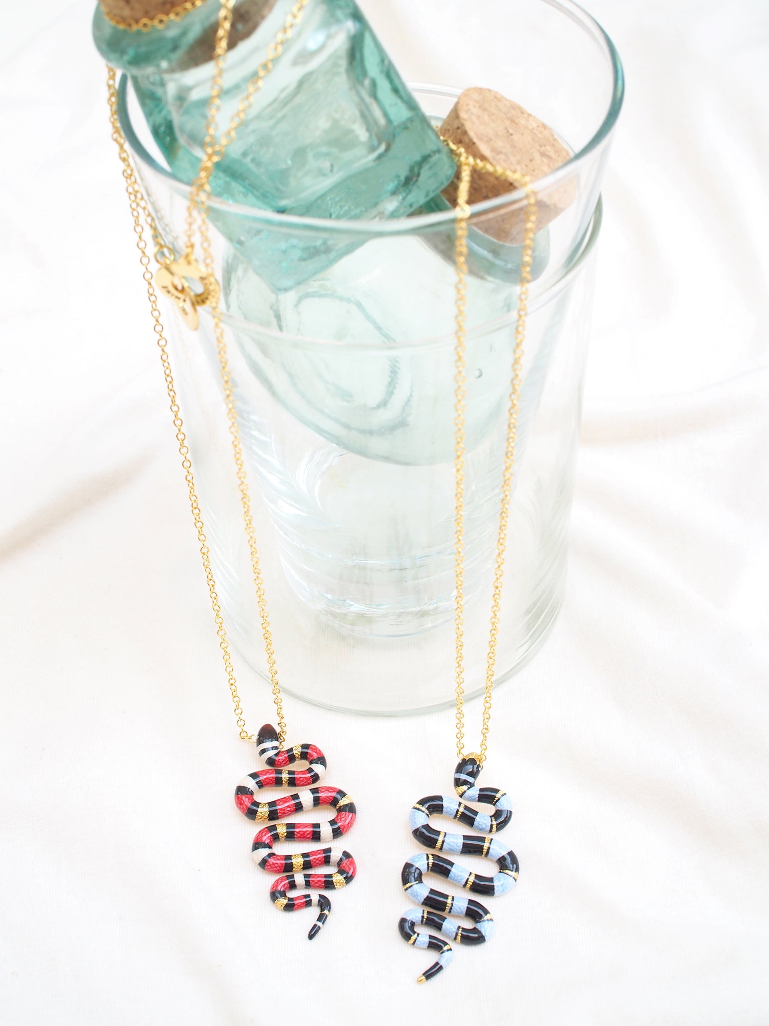 Two milk snake necklaces in red and black, and blue and black, hanging next to a glass bottle.