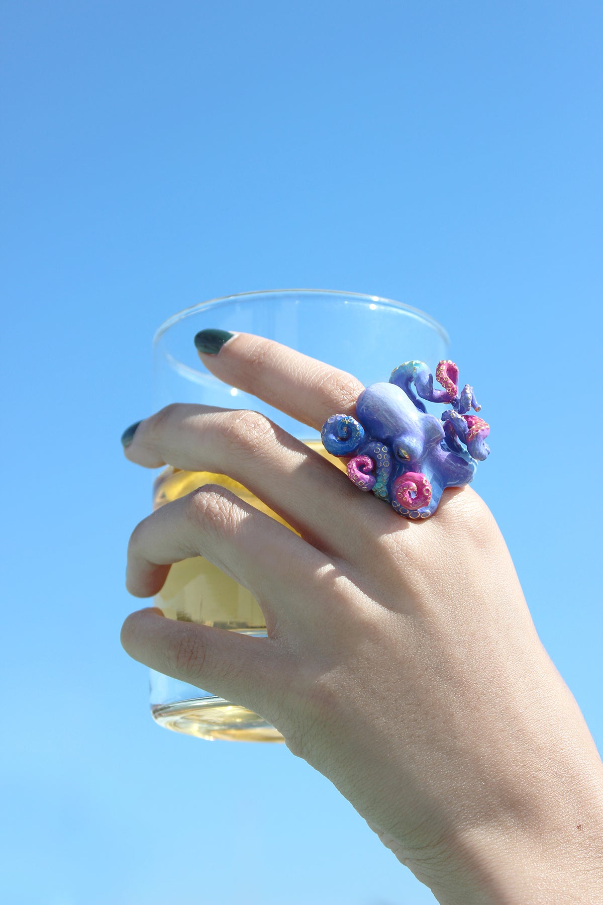 A close-up of a hand adored with a unique octopus ring, holding a glass of water with the sky background.