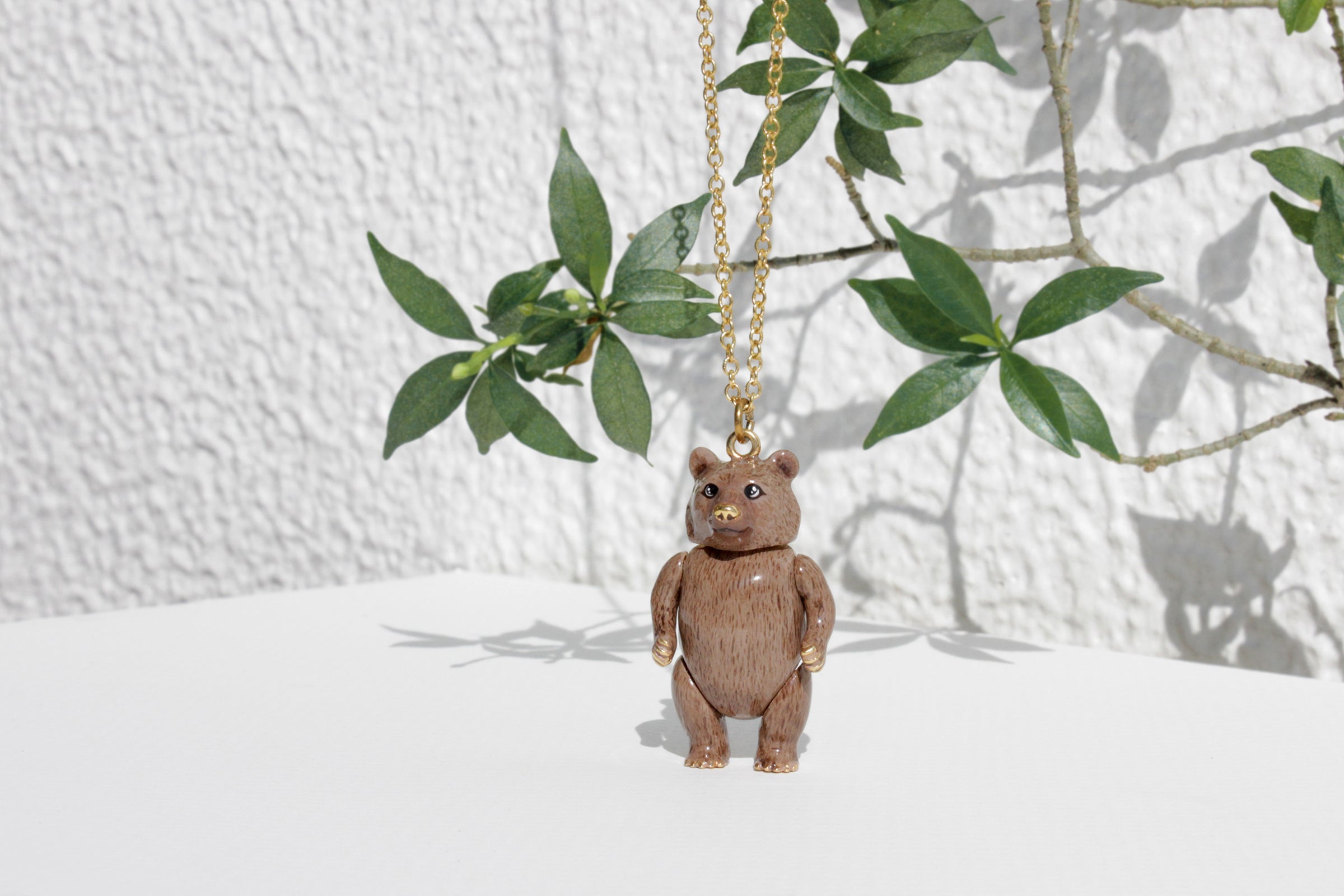 A cute grizzly bear pendant hangs from a gold chain, casting green leaves and shadow on a white surface against a textured wall.