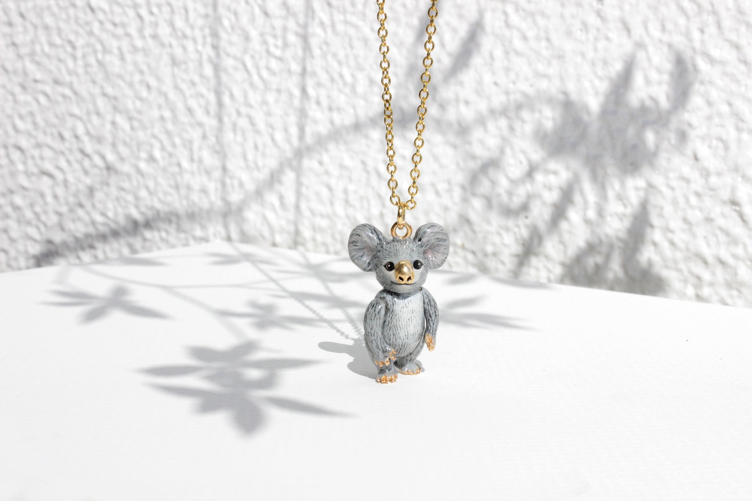 A cute grey koala pendant hangs from a gold chain, casting a delicate shadow on a white surface against a textured wall.