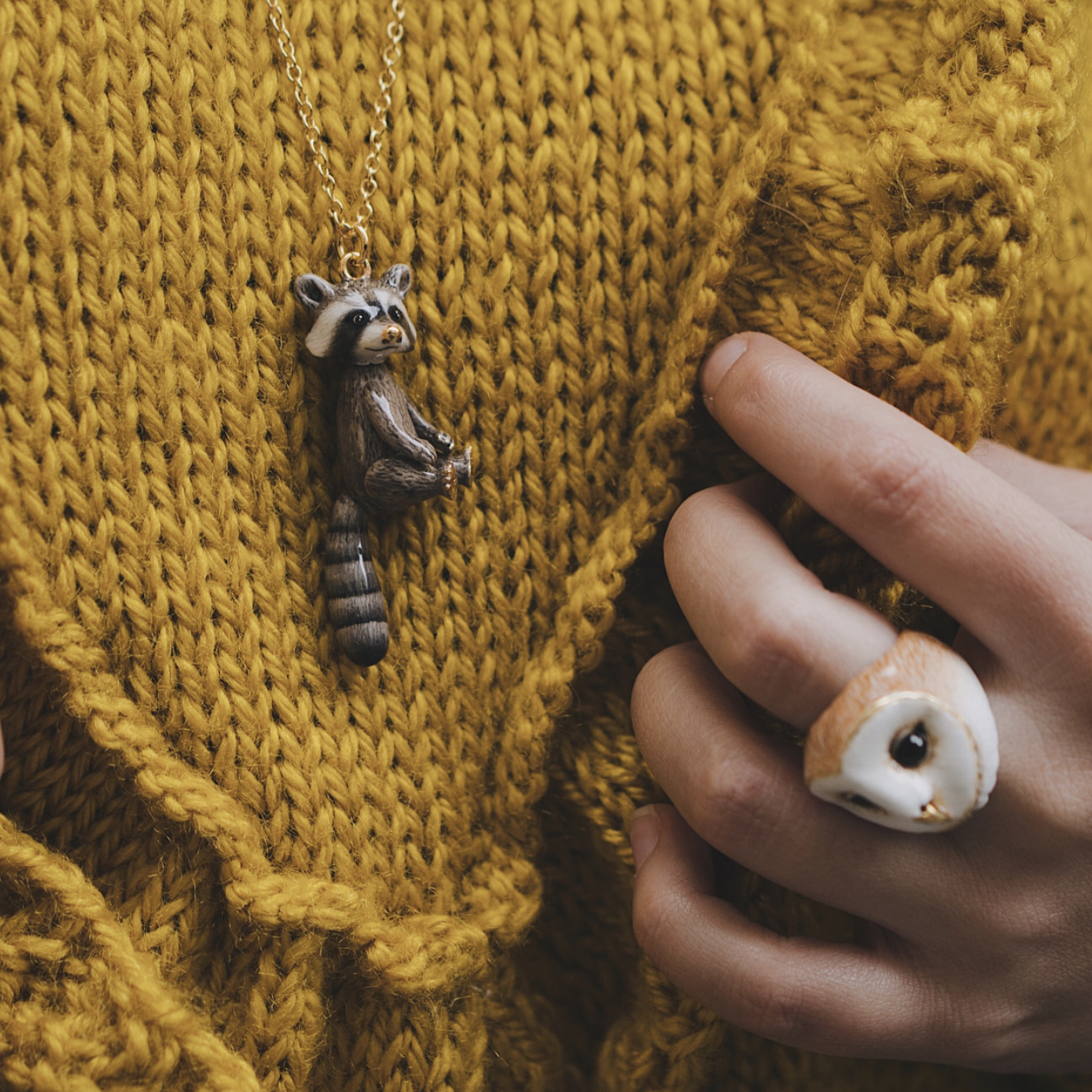 A person wearing a raccoon pendant necklace and an owl ring with a cozy yellow sweater.