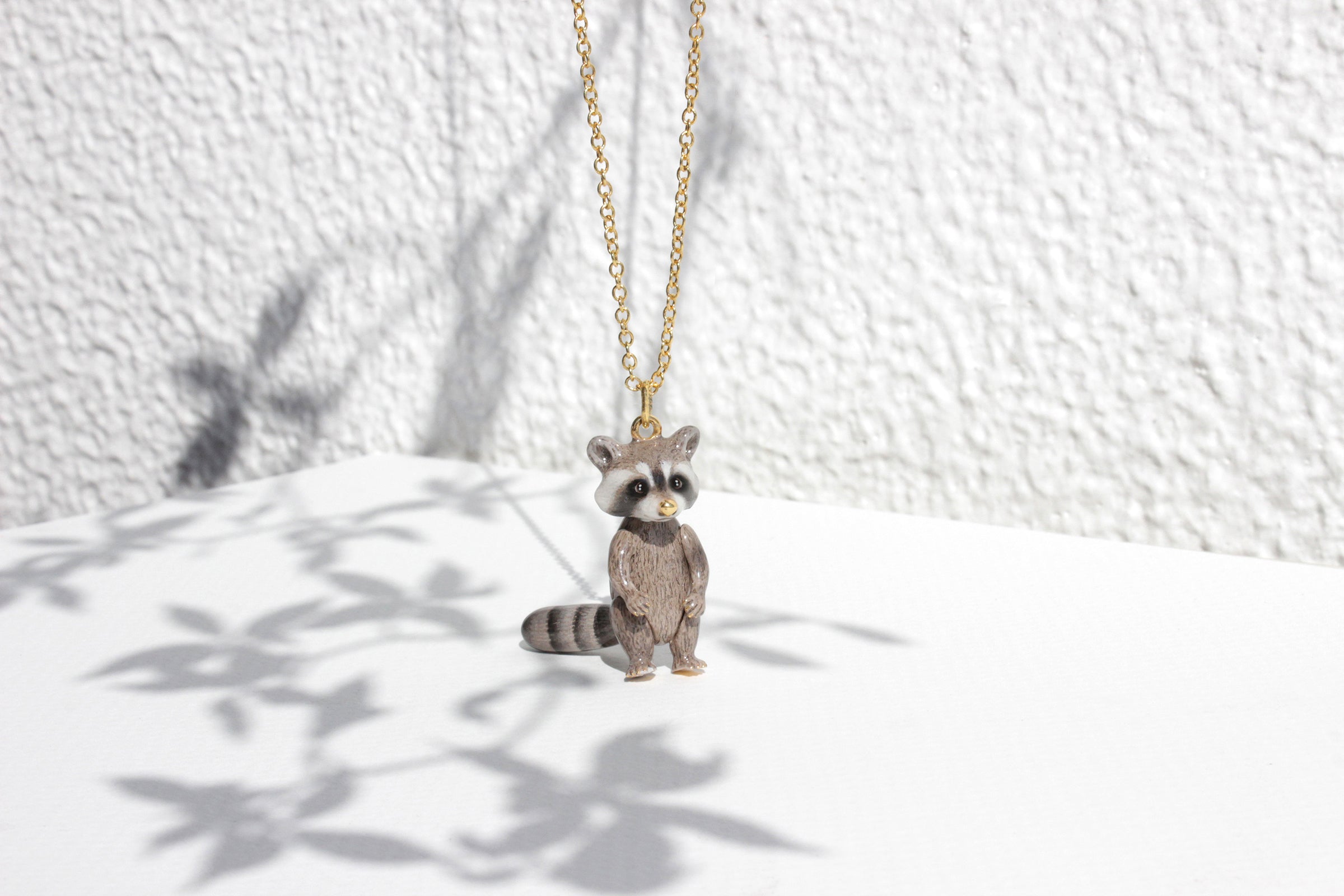 Adorable raccoon pendant necklace set in the white background with the shadow of leaves.