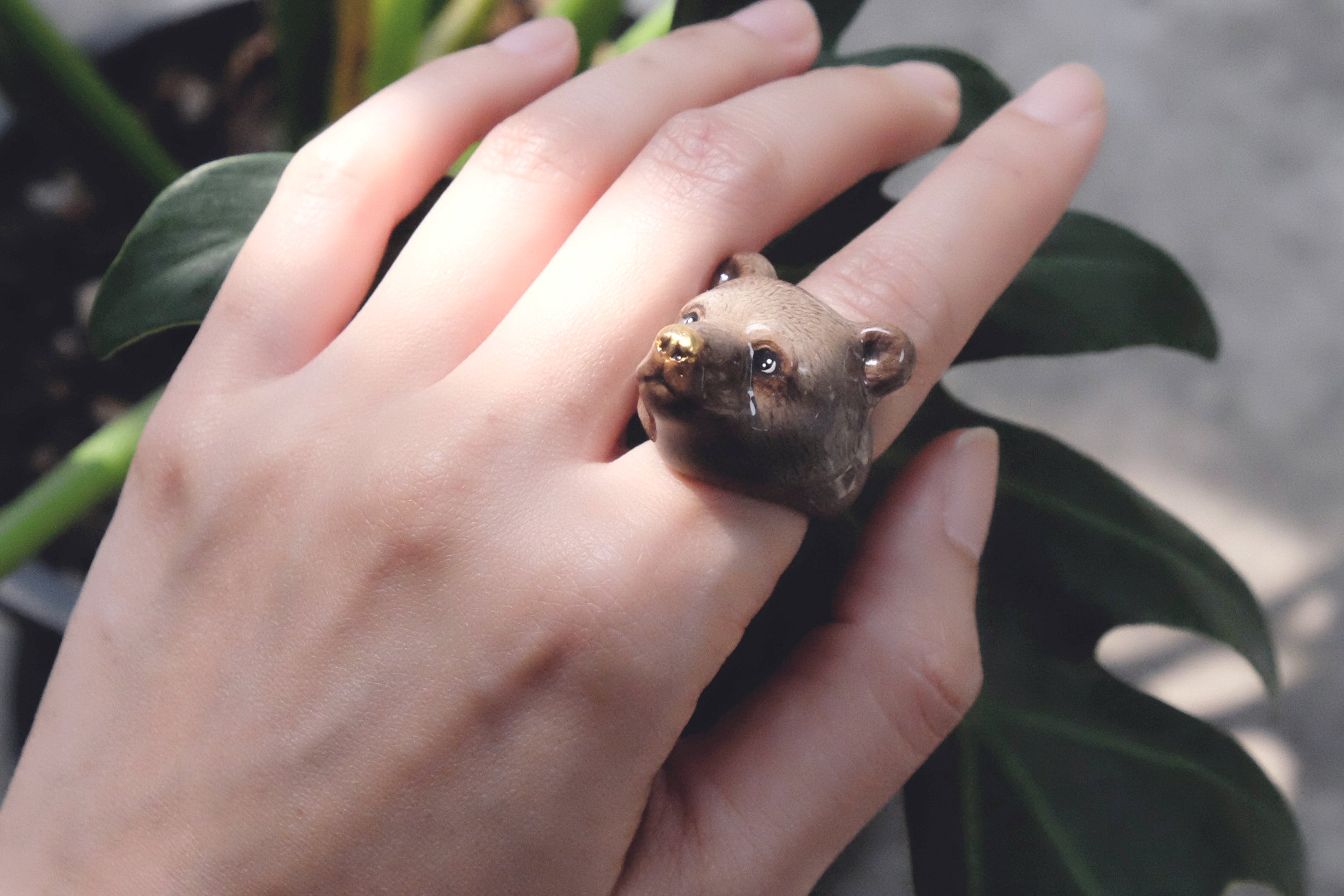 Close-up of a hand adorned with a cute grizzly bear ring surrounded by greenery.