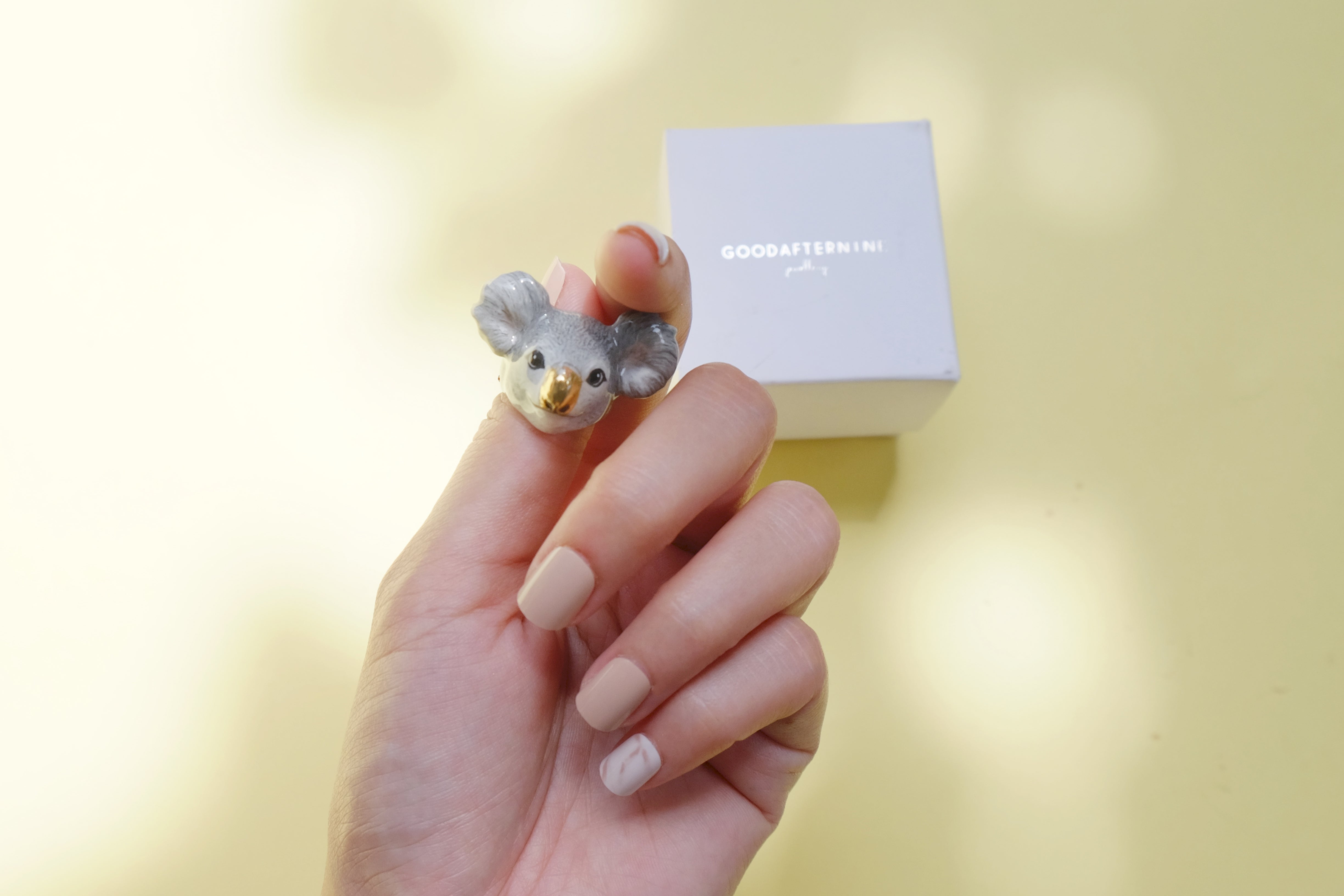 A hand holding a cute koala ring with a smiling face, against a soft yellow background and a nearby white box.