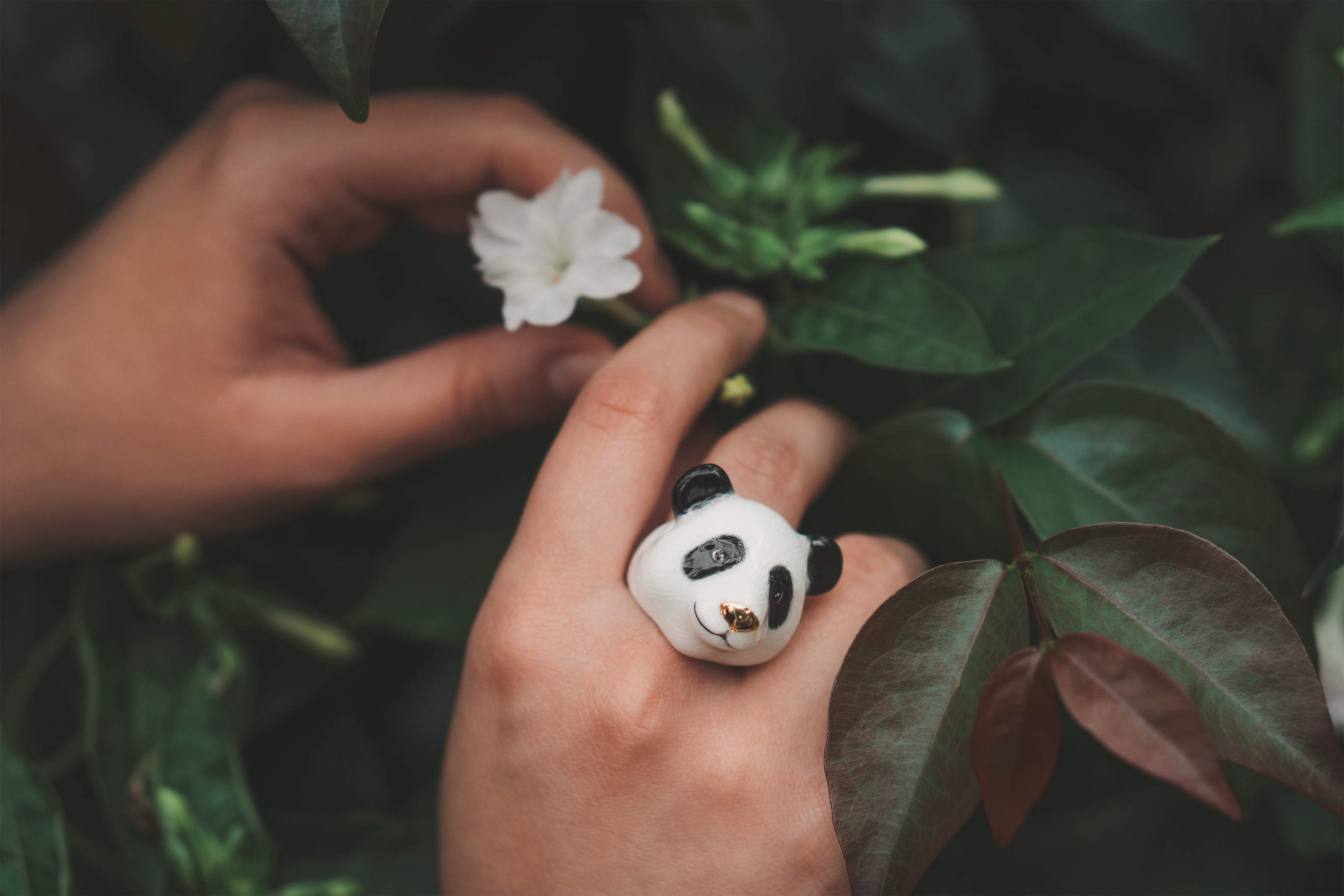 Close up of a hand adorned with a panda ring surrounded by greenery.