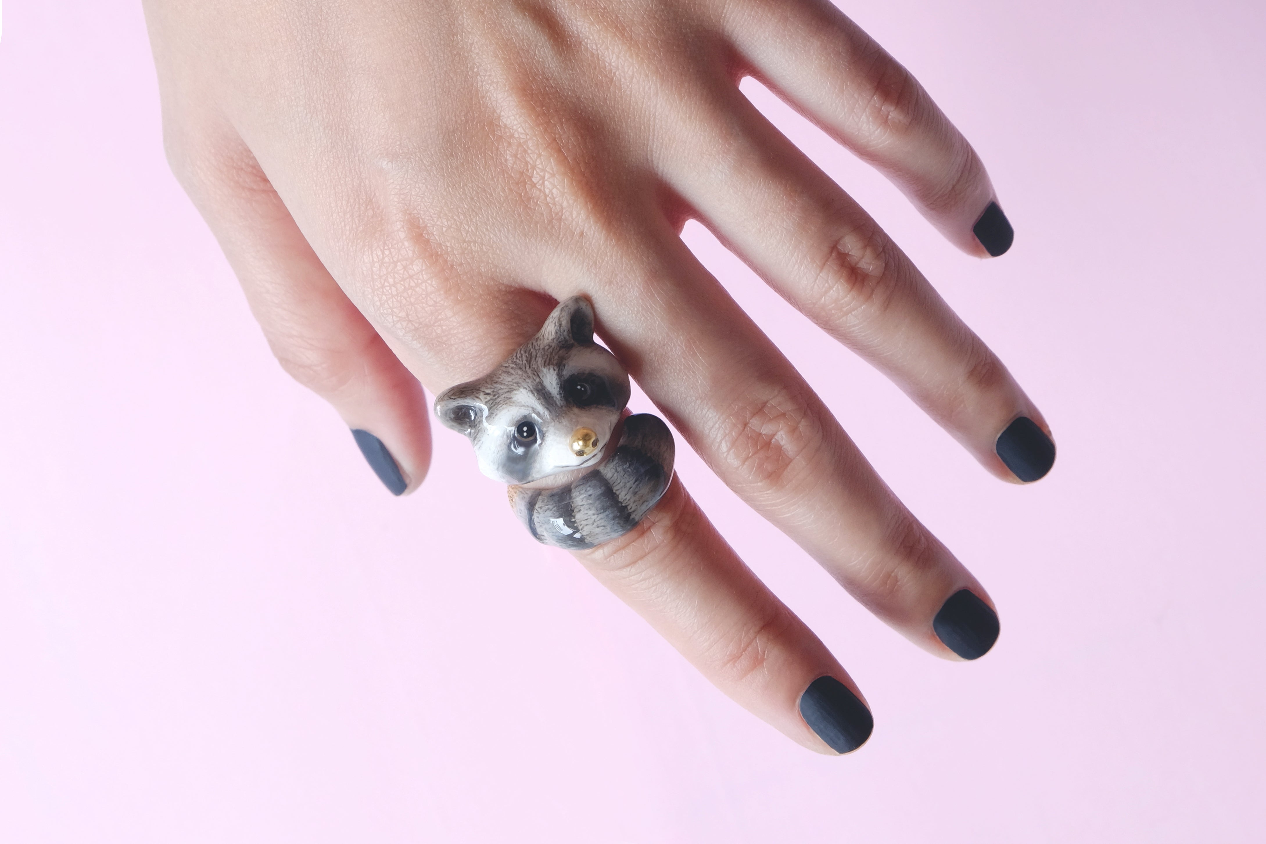 Close-up of a hand adorned with an adorable raccoon ring in a light pink background.