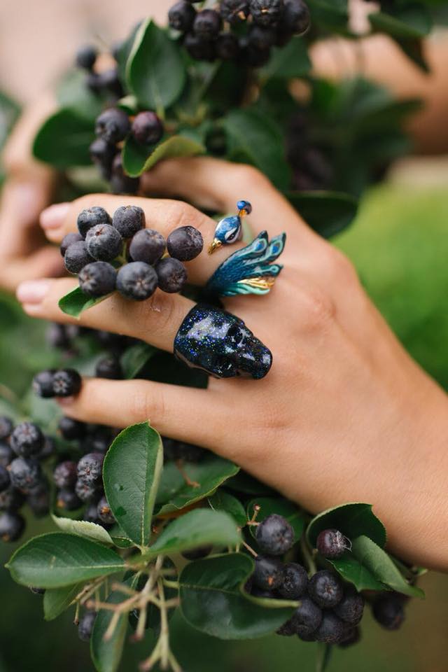  A close-up of a hand adorned with a skull ring and a peacock ring and holding berries in a lush green