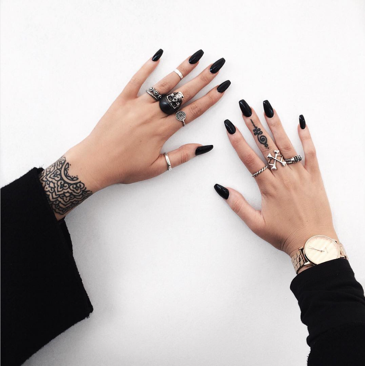 Black manicured nails and unique rings on two hands, one with a tattoo, resting on a white surface.