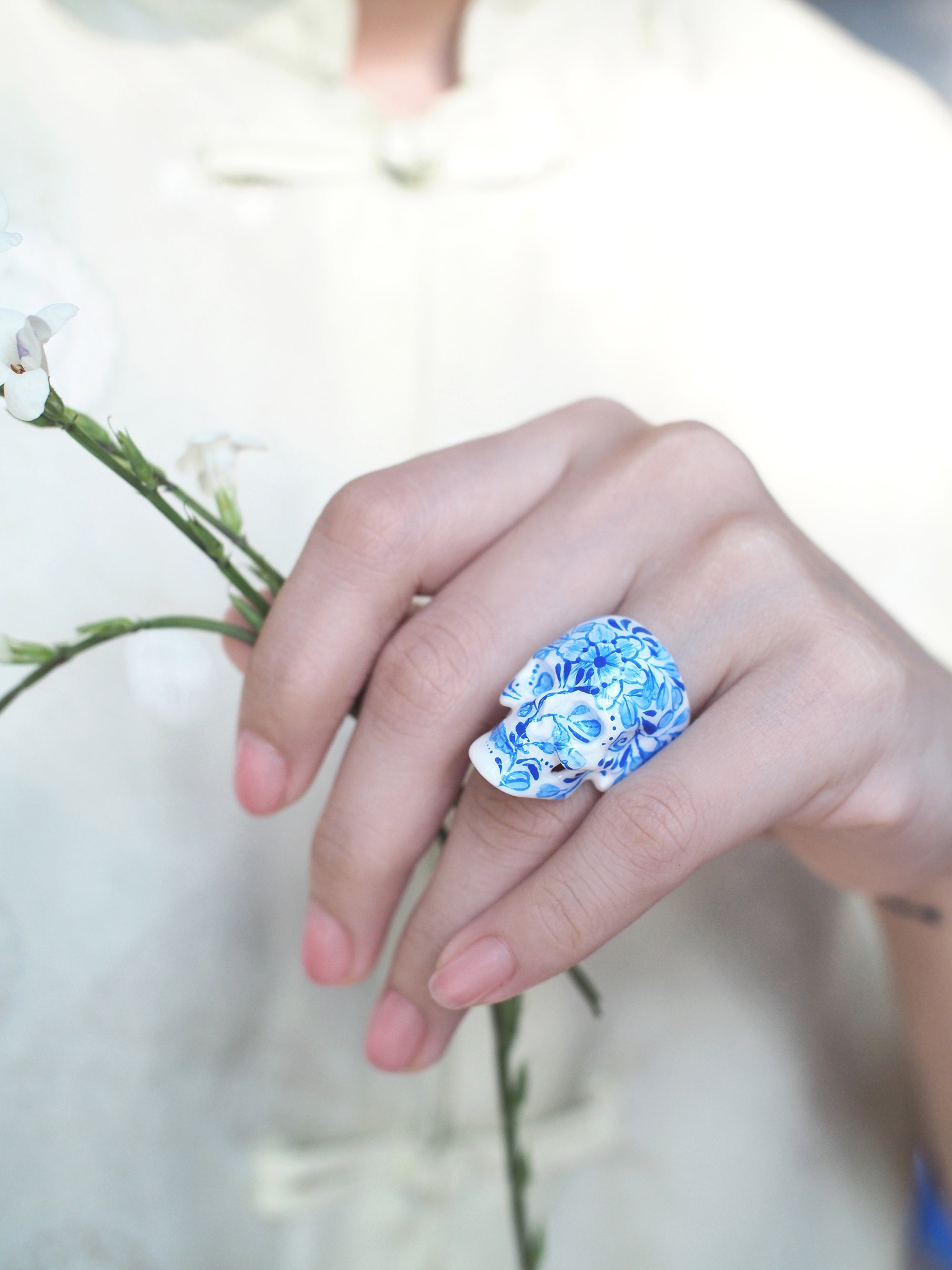 Close-up of a hand holding a flower while wearing a skull ring with artistic blue floral artwork.