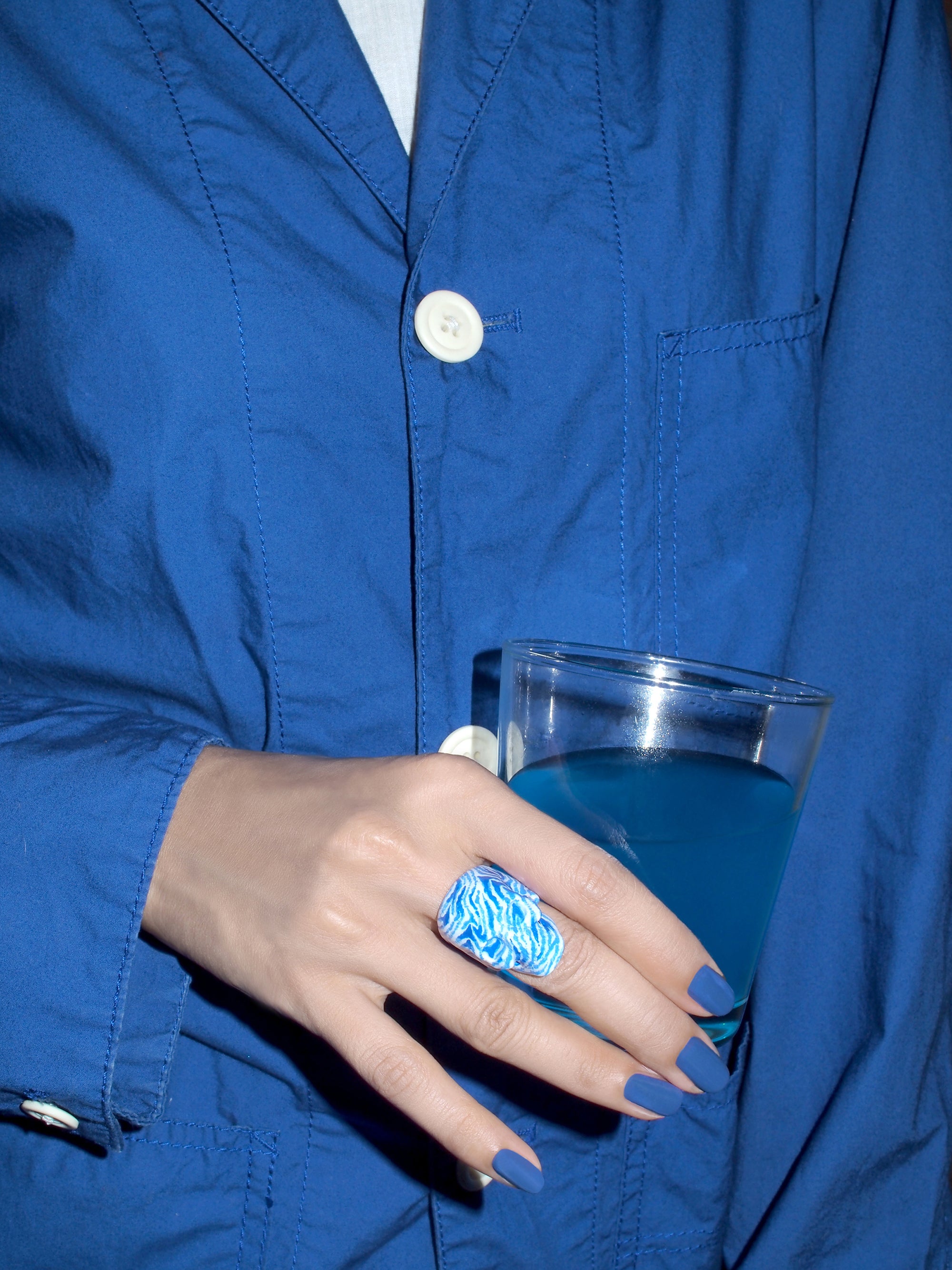 Close-up of a hand in a blue jacket showcasing a white and blue skull ring and a blue drink.