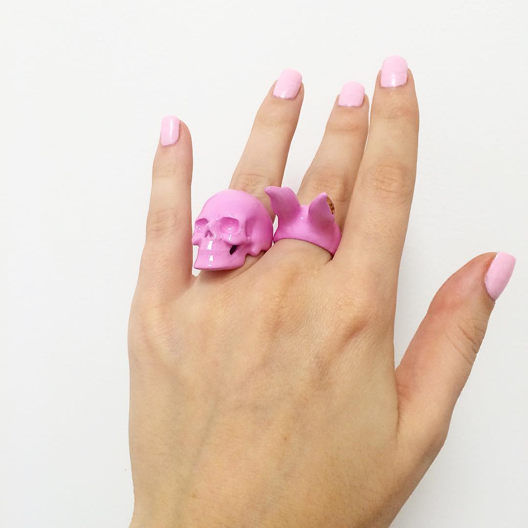 A close-up of a hand with soft pink nail polish wearing a pink skull ring and pink rabbit's ears ring