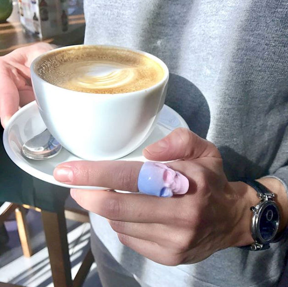 A close-up of a skull ring with a gradient from blue to pink worn on a hand holding a cup of coffee.