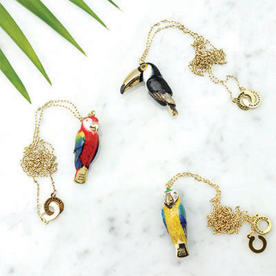 A collection of colorful bird whistle necklaces featuring a parrot, toucan, and macaw on a marble background.