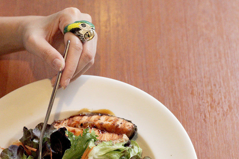 A hand adorned with a blue macaw ring holding a fork above a plate of grilled salmon and salad.