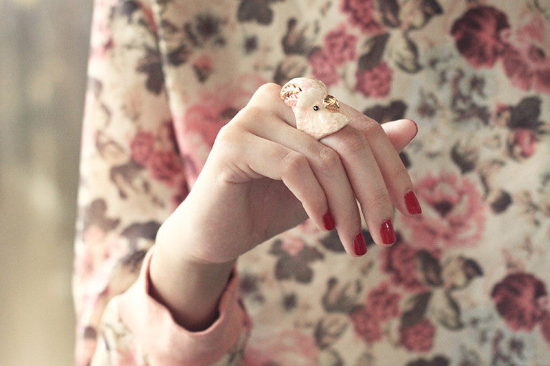 Close up of a hand with red nails wearing cockatoo-shaped ring in soft pink and gold tones.