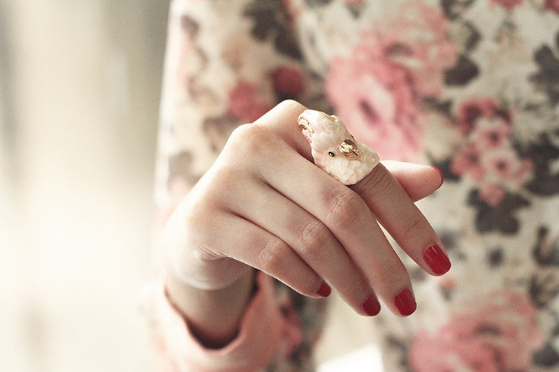 Close up of a hand with red nails wearing cockatoo-shaped ring in soft pink and gold tones.