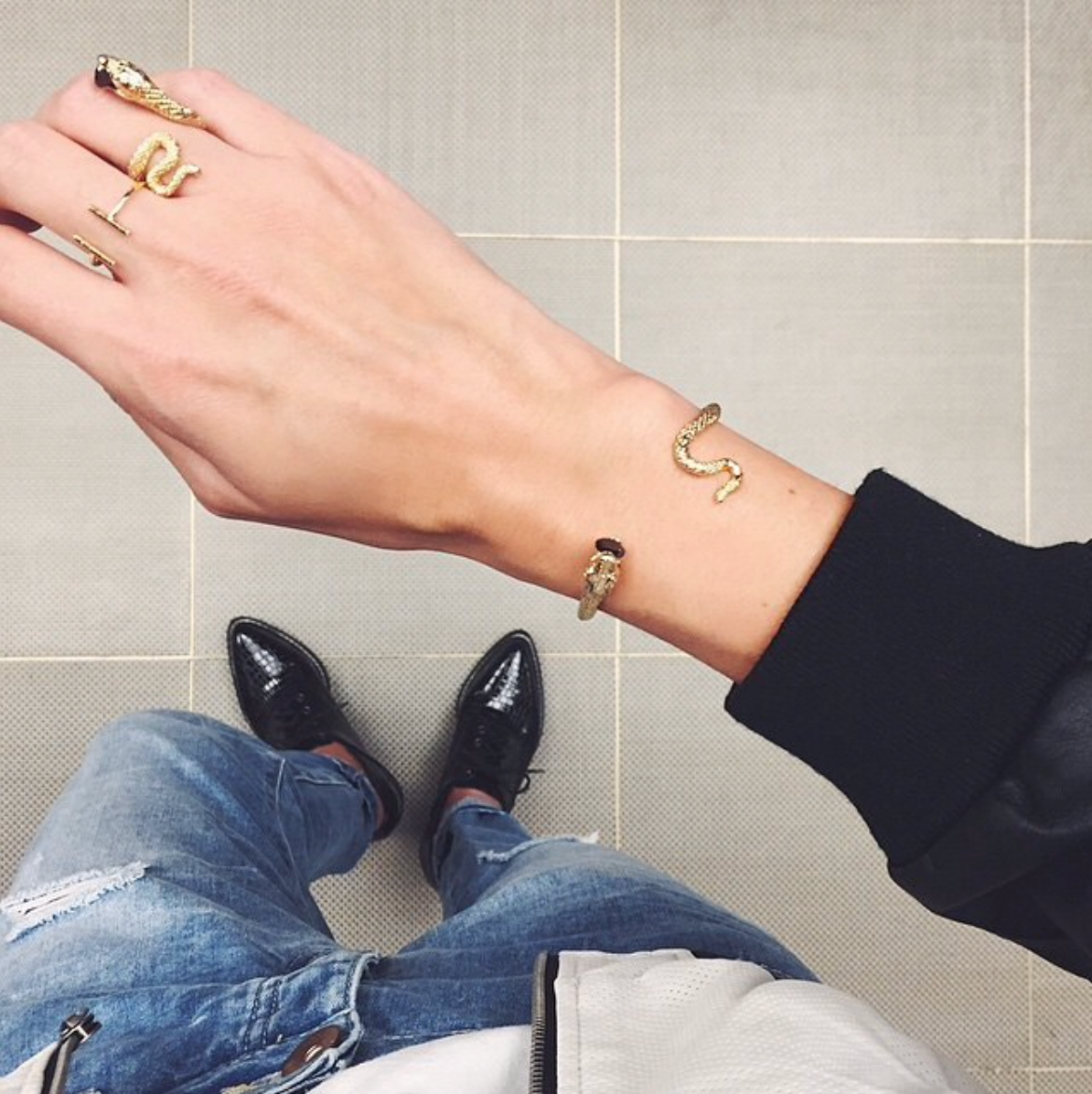 A person wearing a gold sanke jewelry on a person's hand, including rings and a bangle in a casual outfit.