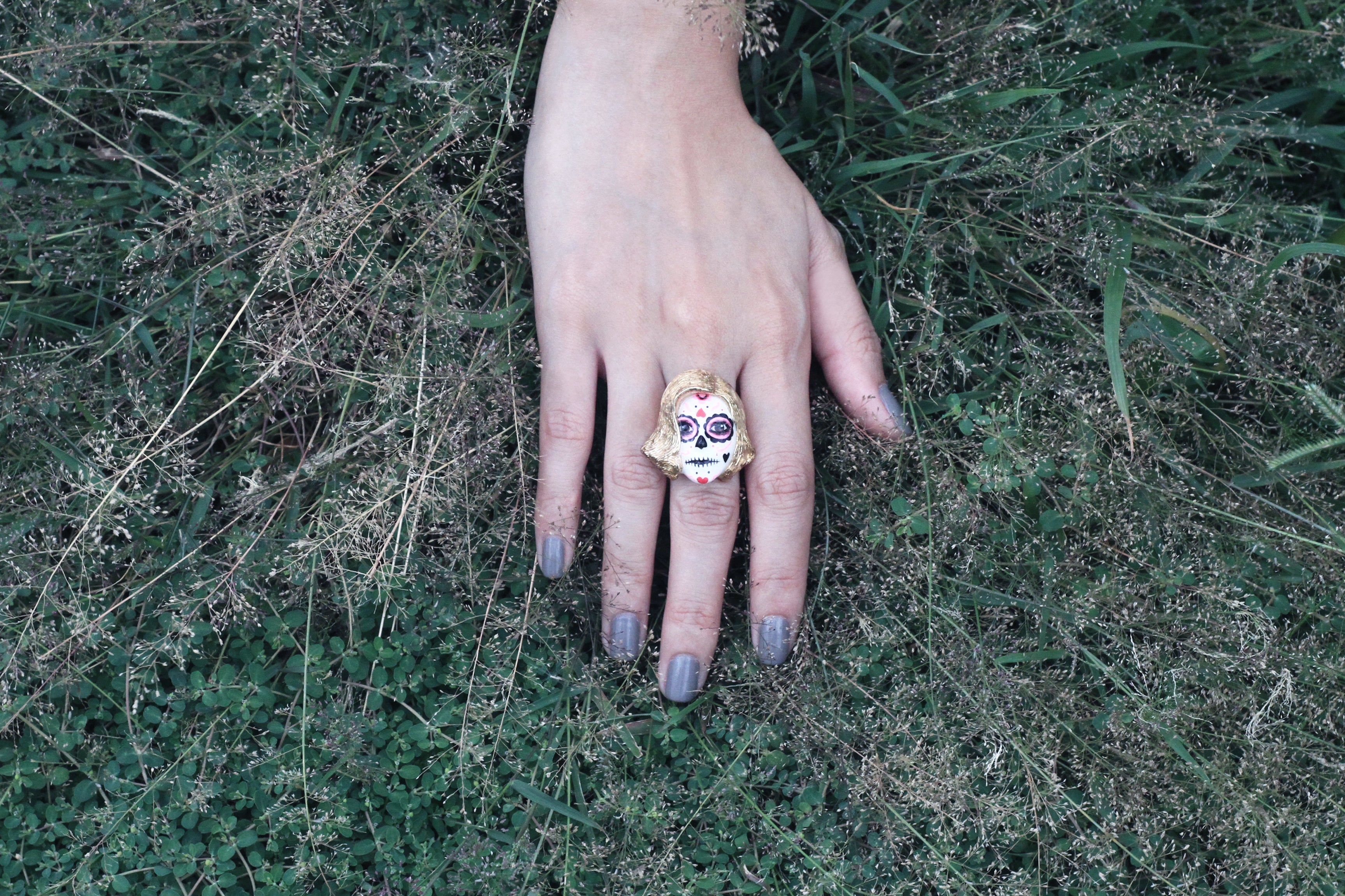 A hand with manicured nails rests on grass, wearing a gold and colorful skull ring, surrounded by greenery.