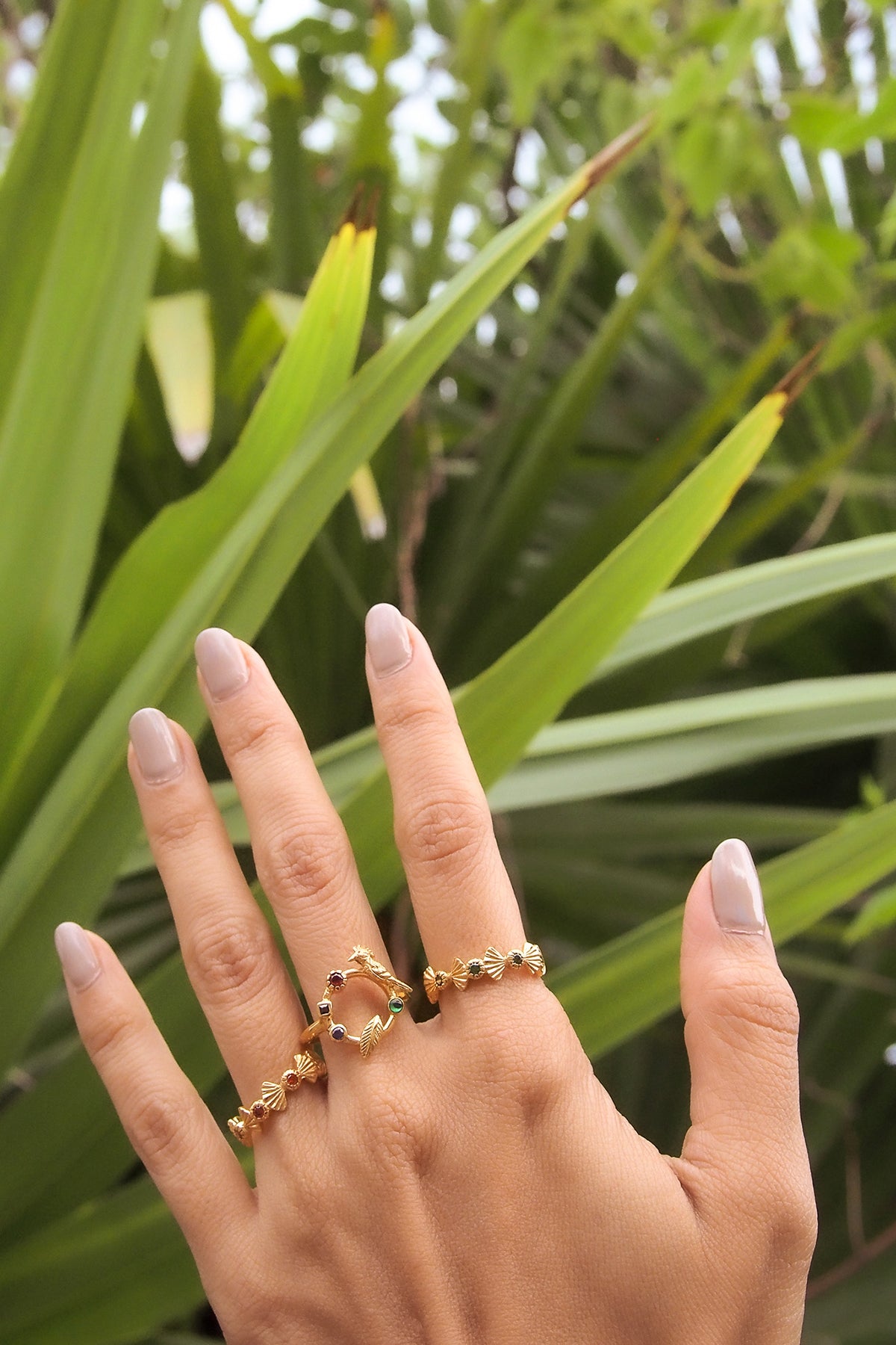 Nature-inspired ring designs displayed on a hand, featuring intricate gold details and vibrant stones amidst a lush green background.