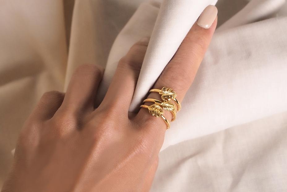 Close-up of a hand wearing three stylish gold rings showcasing beautiful leaf designs on a soft fabric background.