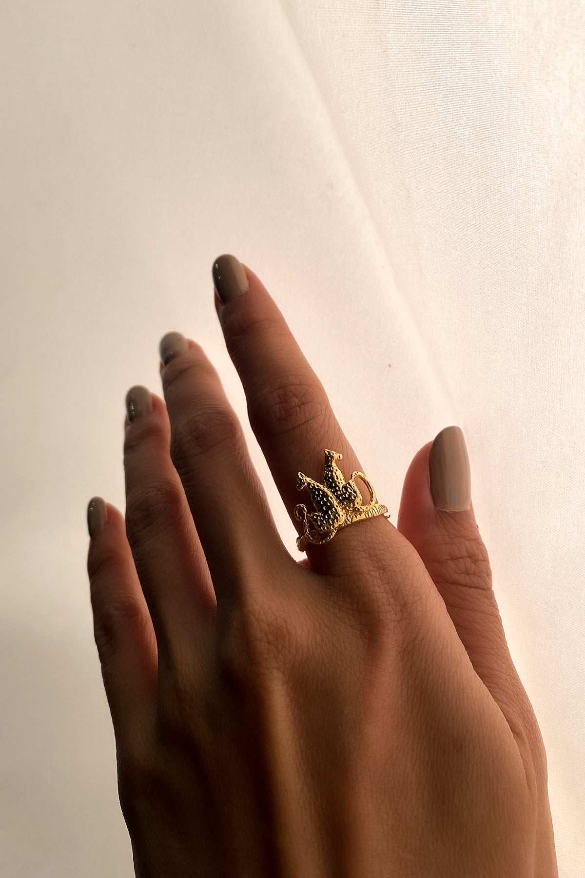 A hand with polished nails wearing a gold leopard ring against a soft, light background, highlighting the ring's intricate details.