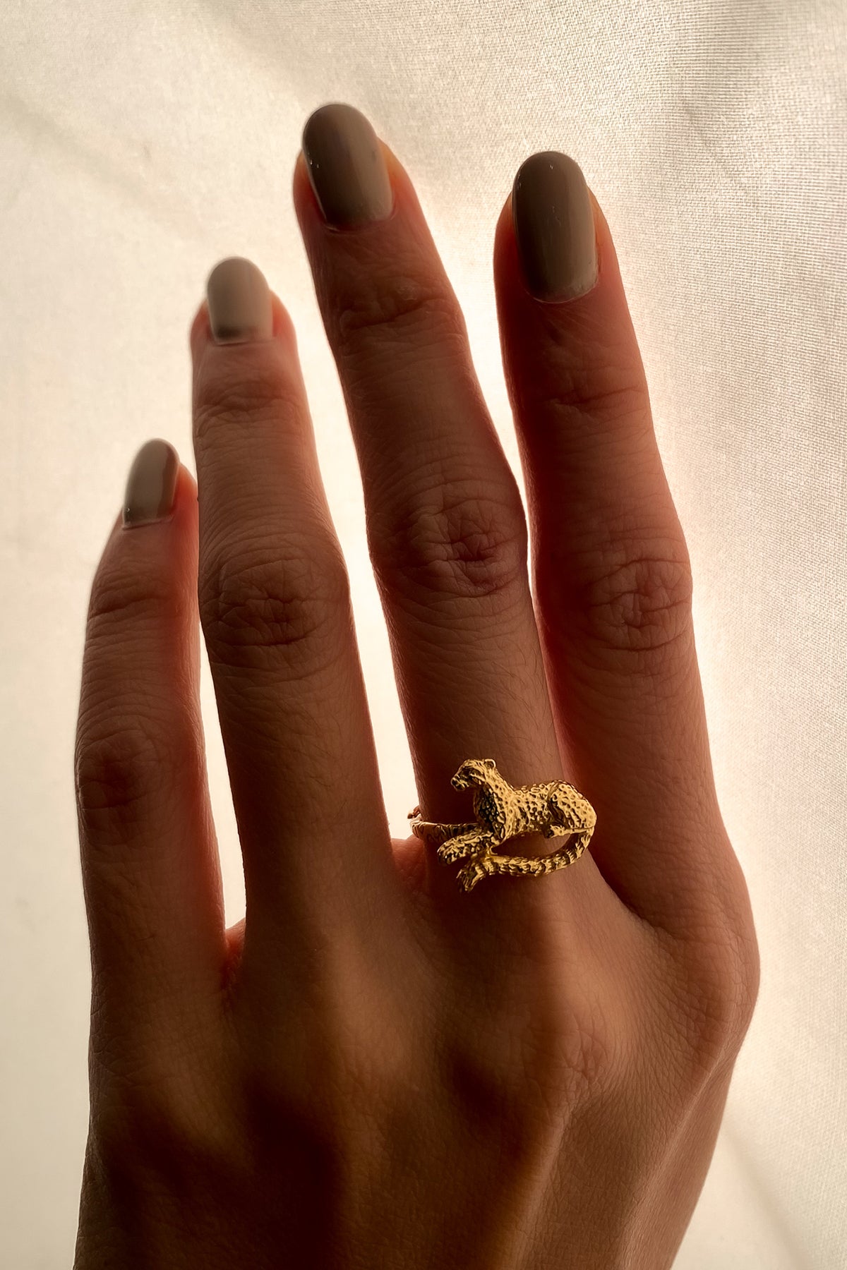 A hand displaying a gold ring featuring leopard design, with neutral-toned nails against a softly lit background.