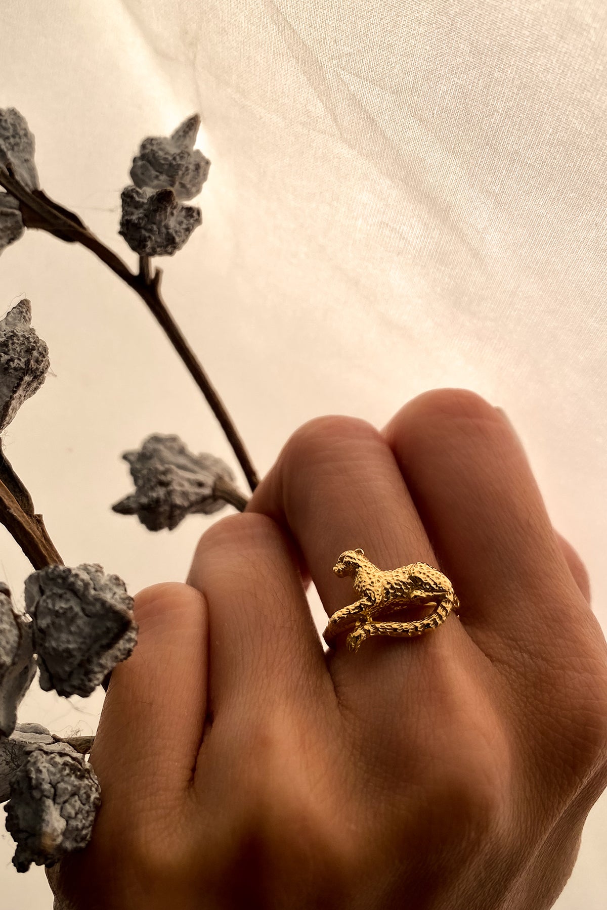 A hand adorned with polished nails wearing a gold leopard ring against a soft, light background, highlighting the ring's intricate details.